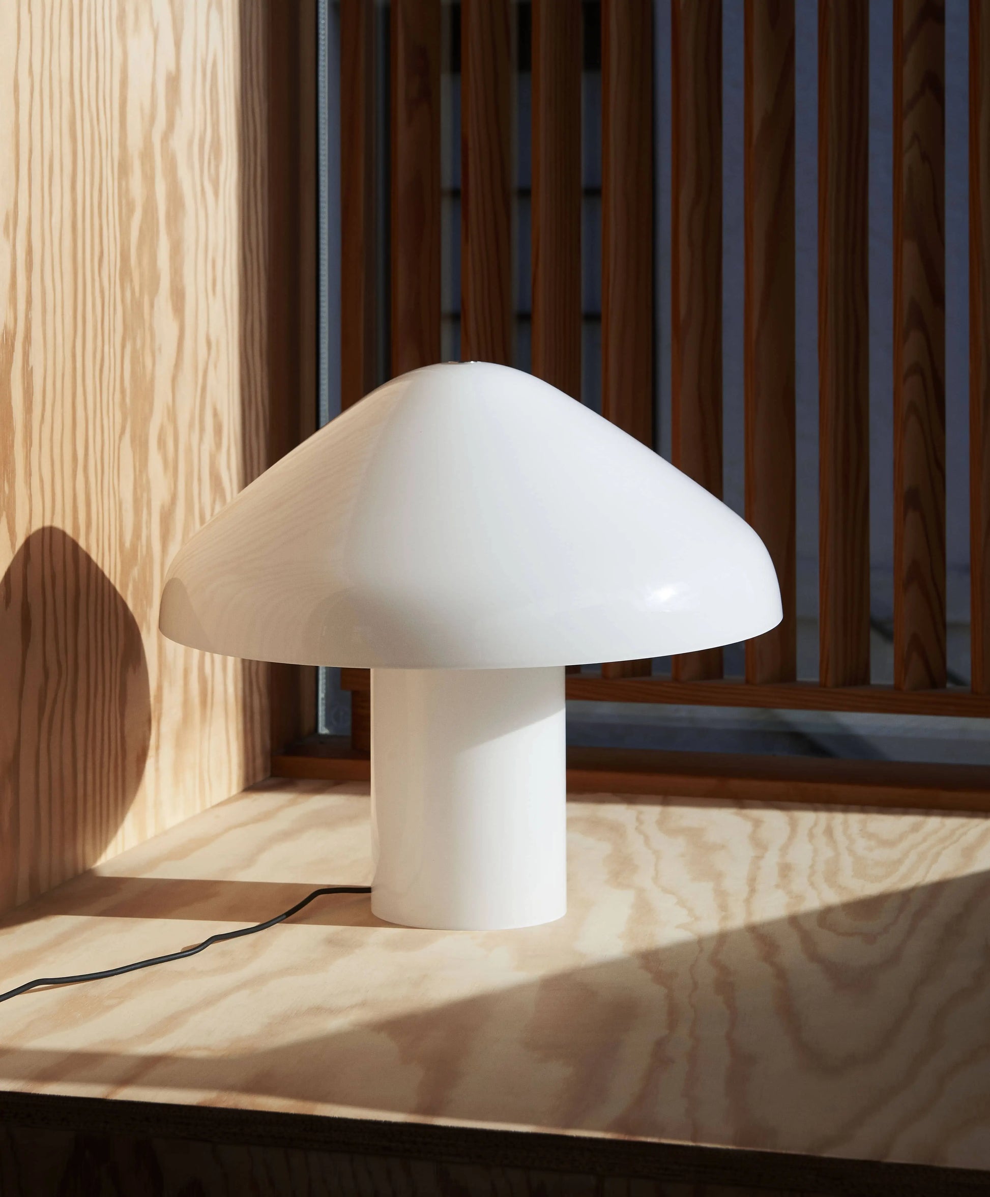 The HAY Pao Portable Lamp by HAY, with its modern white mushroom shape and glossy finish, sits on a light wood surface near vertical wooden slats, sunlight highlighting its portable design.