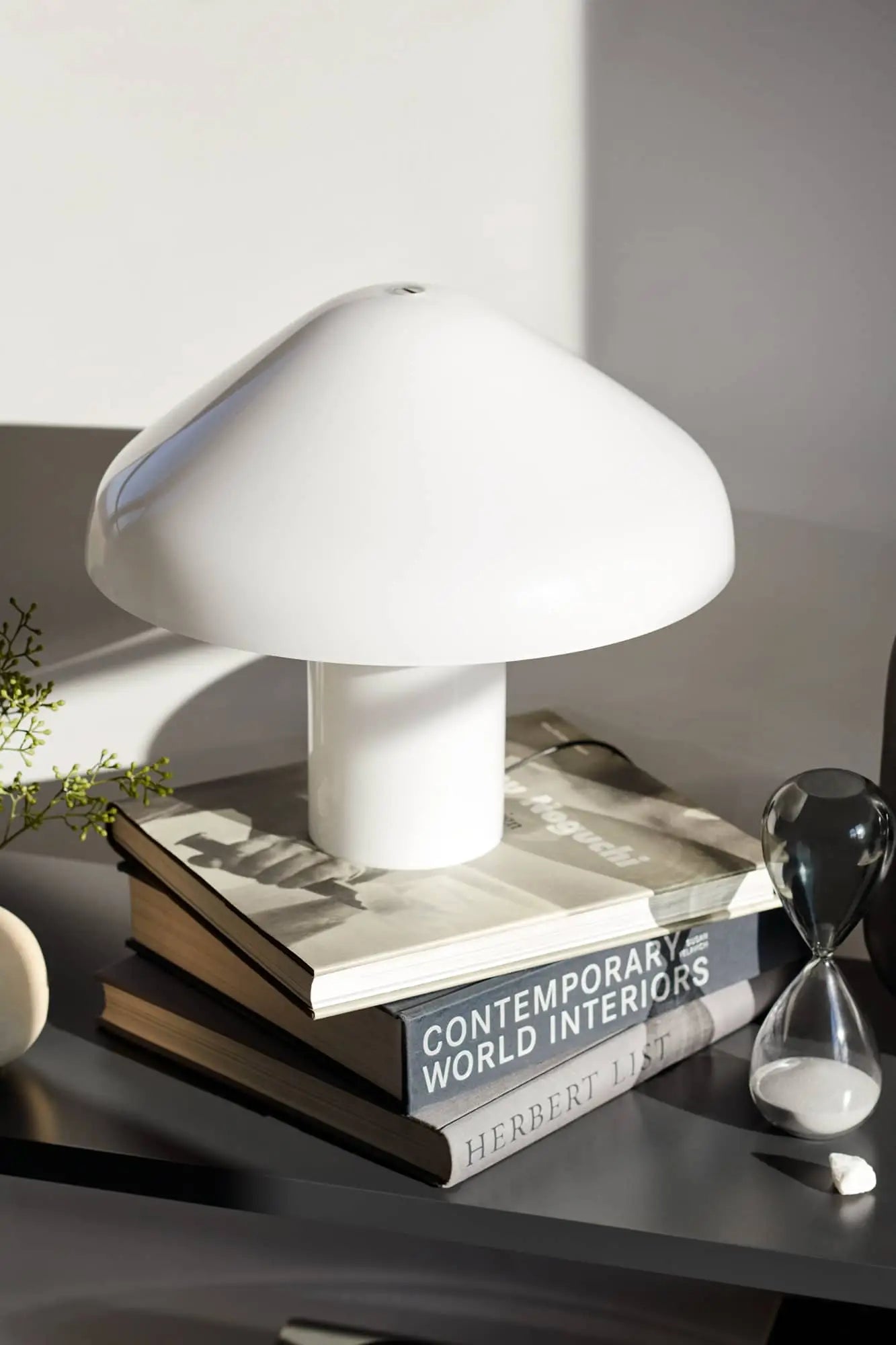 A modern white HAY Pao Portable Lamp by HAY sits atop books—including CONTEMPORARY WORLD INTERIORS and HERBERT LIST—beside a small hourglass and greenery on a dark surface.