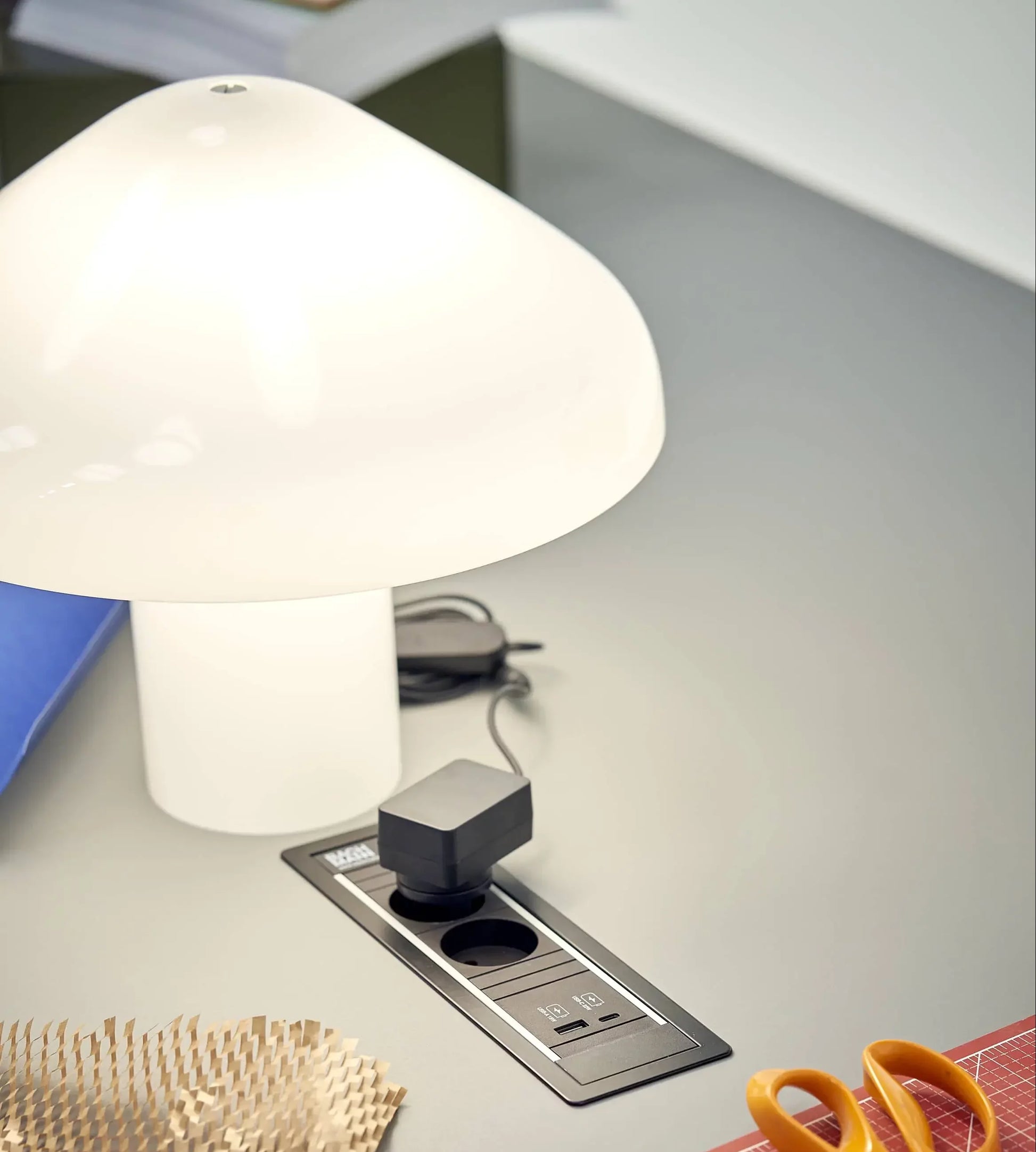 A white HAY Pao Portable Lamp by HAY sits on a gray desk beside a blue folder, a power outlet with a black charger, and part of a yellow object, possibly scissors, in the foreground.