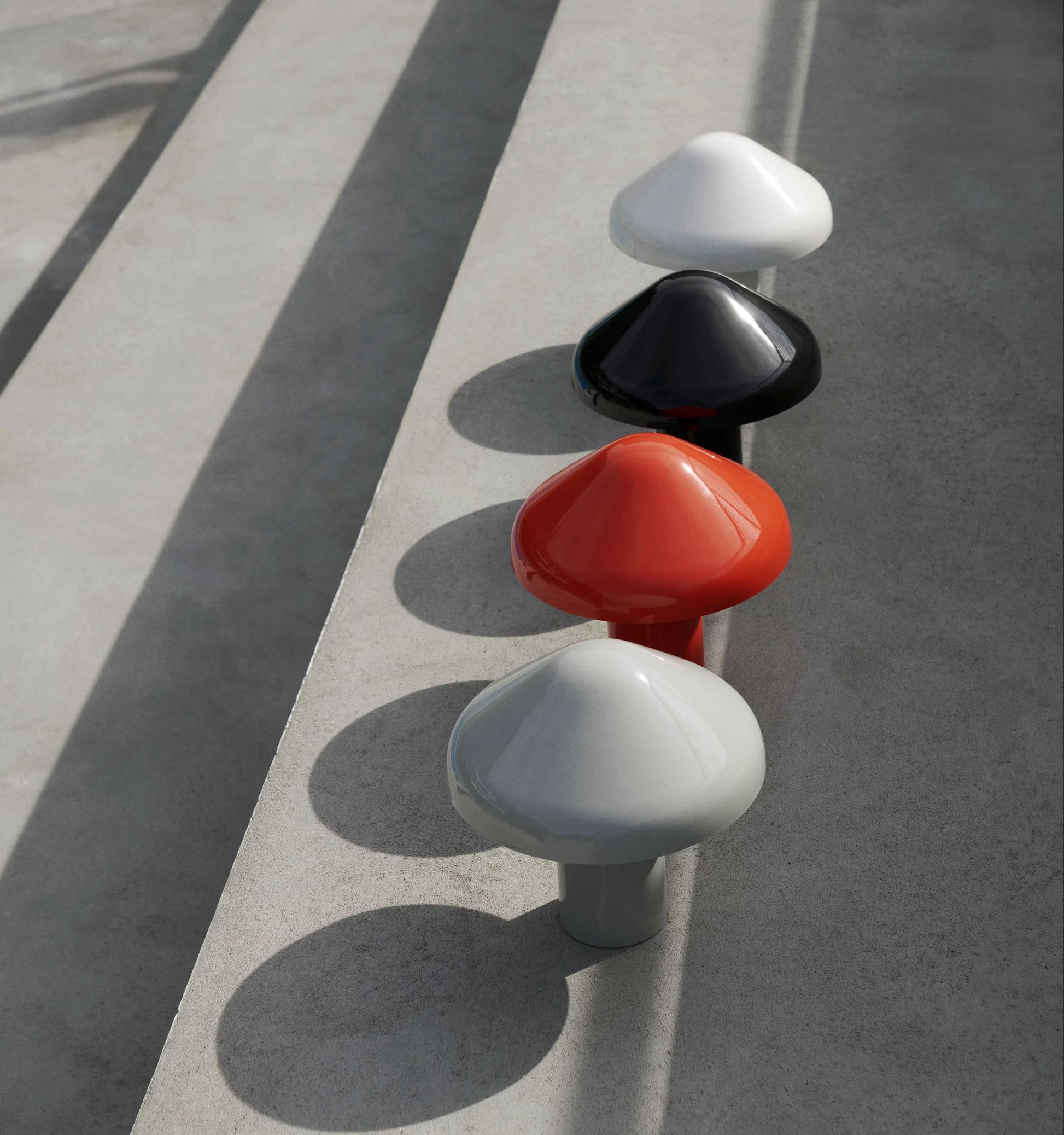 Four HAY Pao Portable Lamps by HAY—grey, orange, black, and white—are lined up on concrete steps in sunlight, casting unique shadows. Their portable design brings both elegance and versatility to any space.