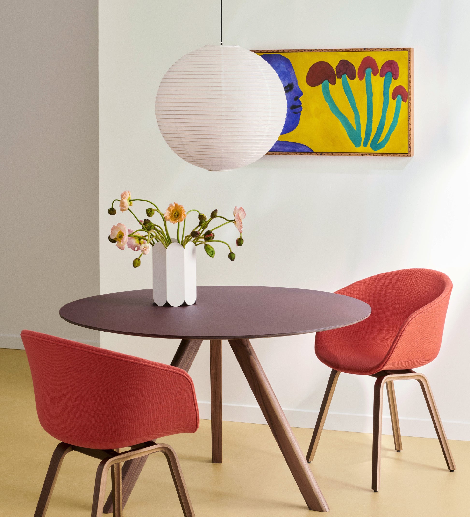 A modern dining area with a round dark table, two red upholstered chairs, a white vase of flowers, the HAY Paper Shade 50cm Pendant Light by HAY overhead, and a colorful wall painting featuring a blue face and red shapes.