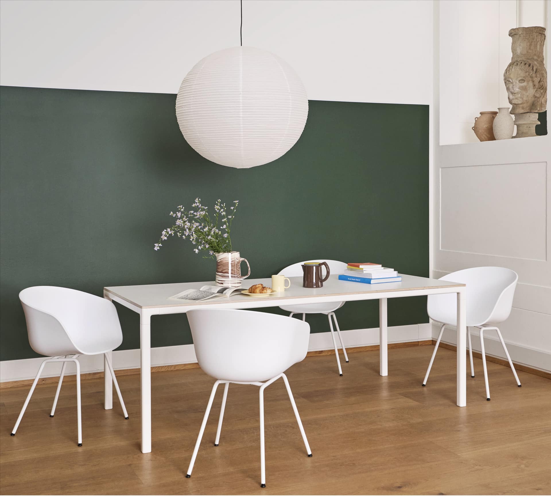 A modern dining area includes a white table, four white chairs, a green accent wall, and the HAY Paper Shade 60cm Dia. With Pendant Cord Set by HAY, LED compatible. Books, mugs, flowers in a vase, and pastries sit on the wooden floor.