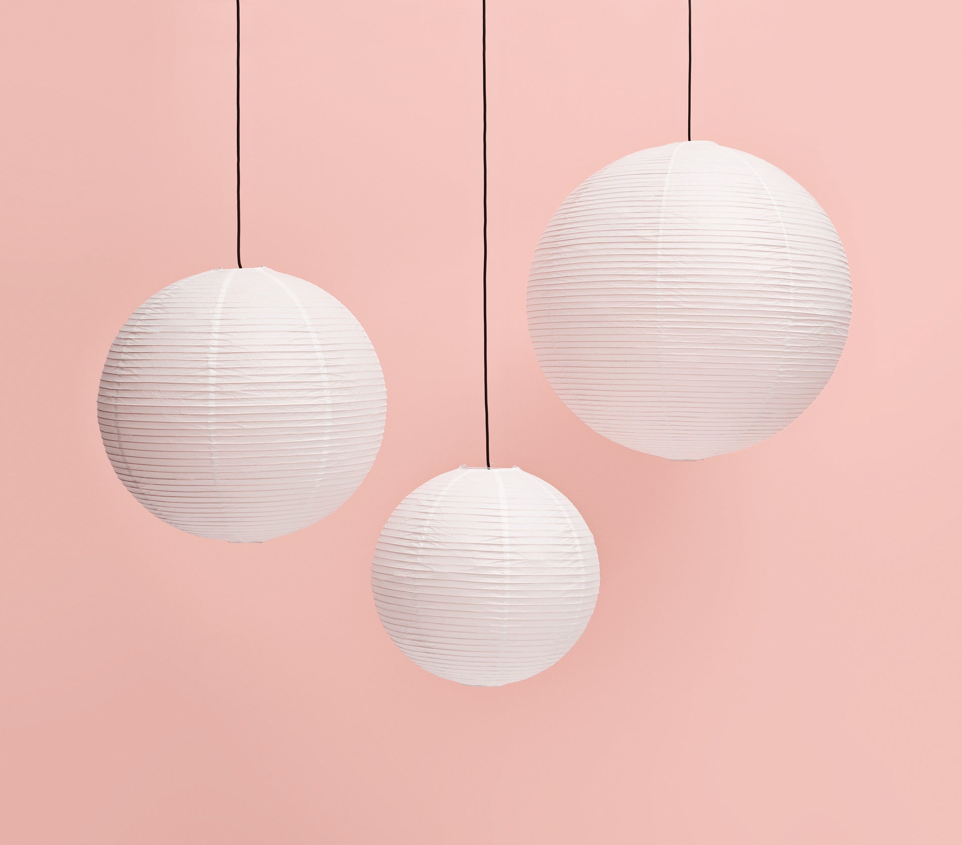 Three HAY Paper Shade 60cm pendant lights with ribbed white rice paper shades and black cords hang against a soft pink background. LED compatible, the minimalist design by HAY creates a simple, elegant composition.