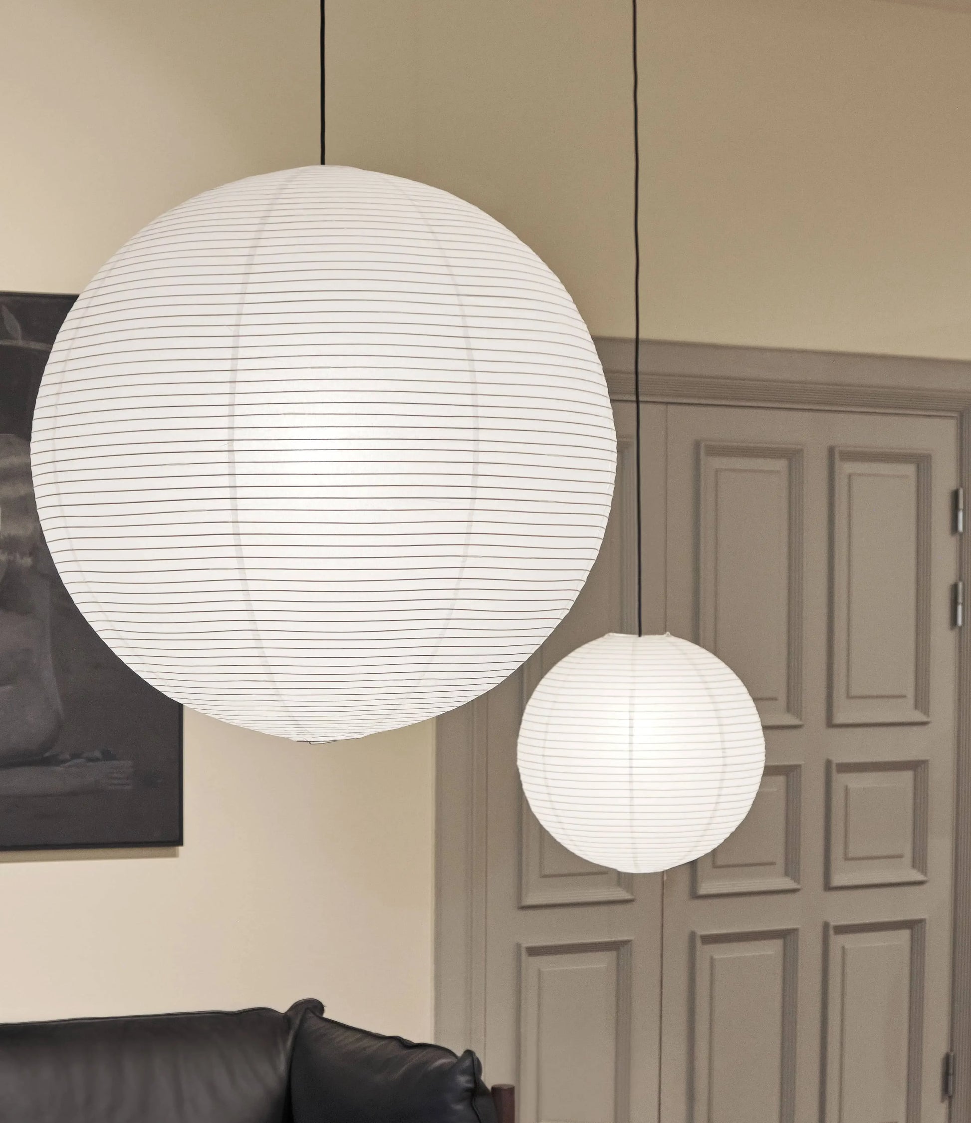 Two HAY Paper Shade 80cm Dia. pendant lights with cord sets hang from the ceiling in a room with beige walls, a closed beige door, and part of a black sofa visible in the foreground.