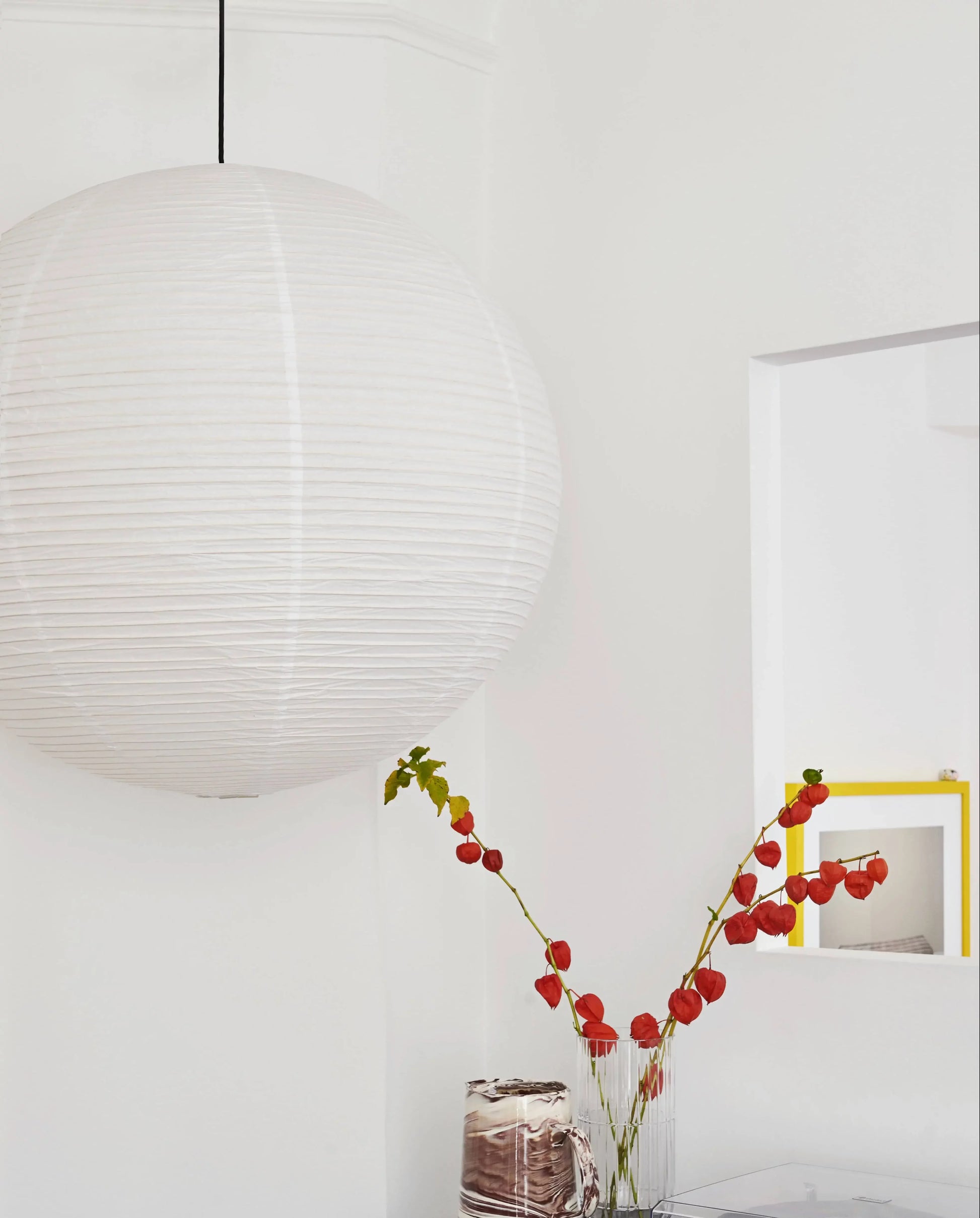 The HAY Paper Shade 80cm Dia. With Pendant Cord Set by HAY hangs from the ceiling near a vase of orange lantern flowers on a table, set against a white wall with a small window and yellow-framed mirror in the background.