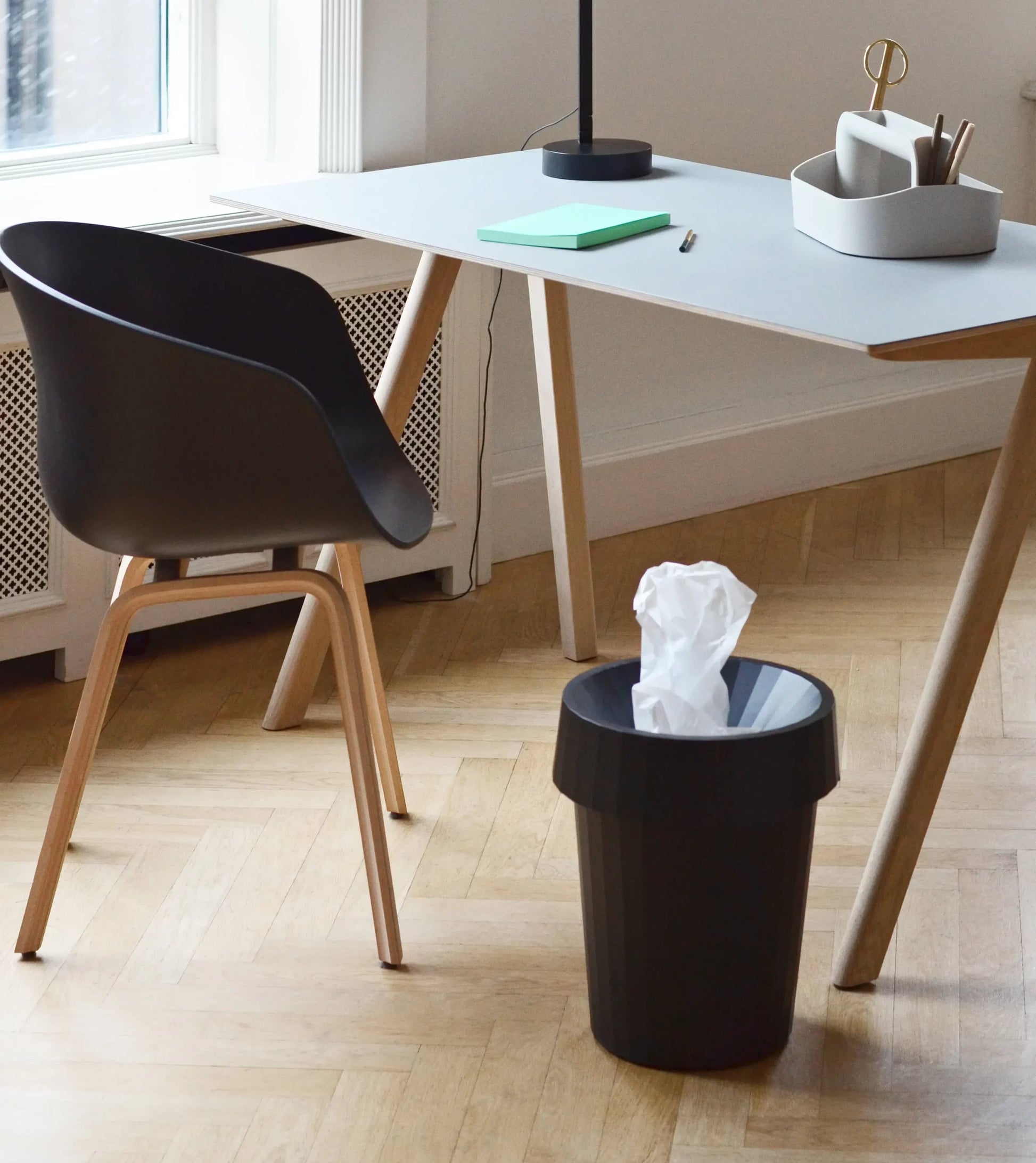 A modern office in muted shades features a black chair, light wooden desk with office supplies and notebook, and a durable HAY Shade Bin by HAY filled with crumpled paper on a light wood floor near a window.