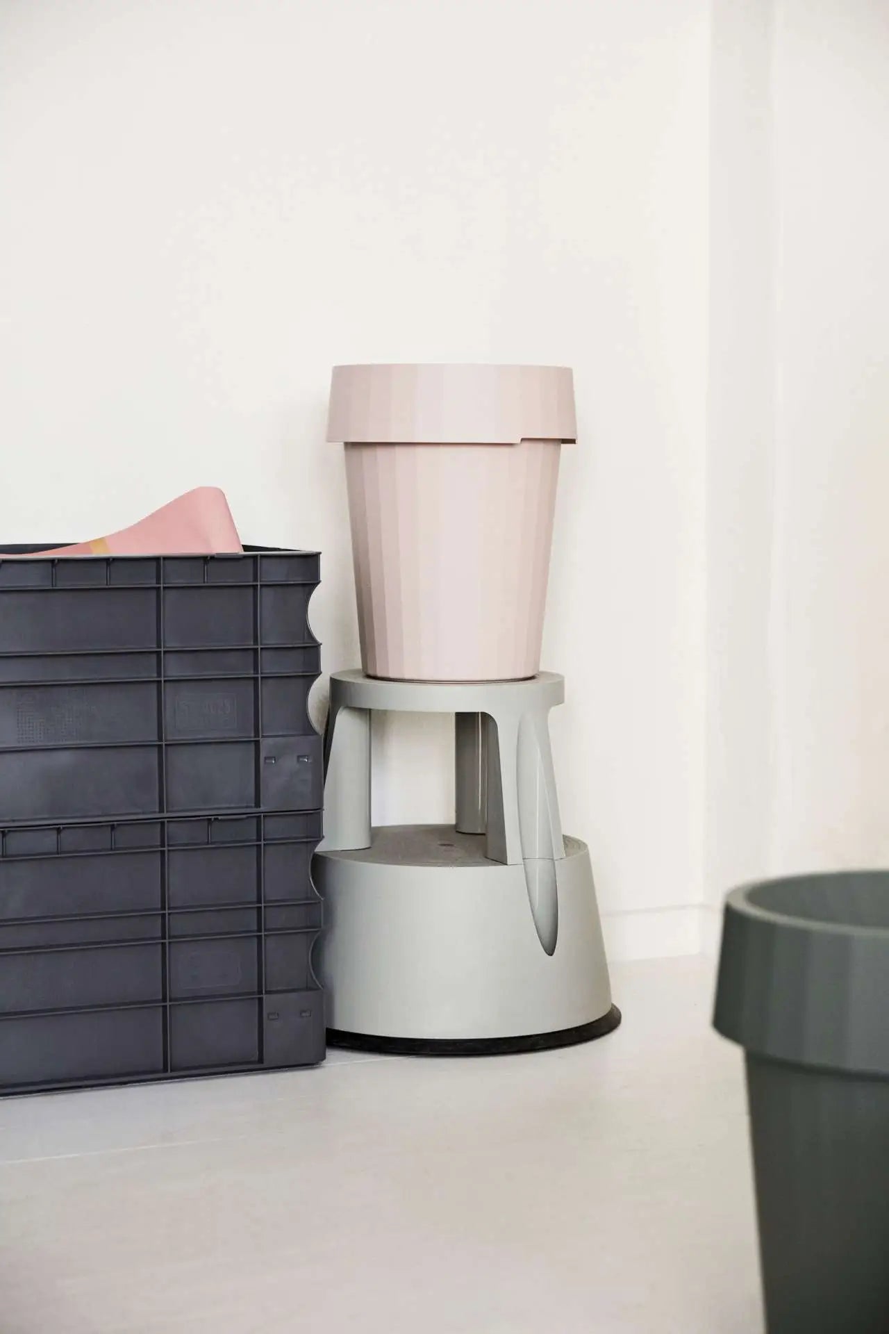 A beige plant pot sits on a light gray step stool beside durable black polypropylene storage containers and the HAY Shade Bin in green, all neatly arranged on a white floor against a white wall.
