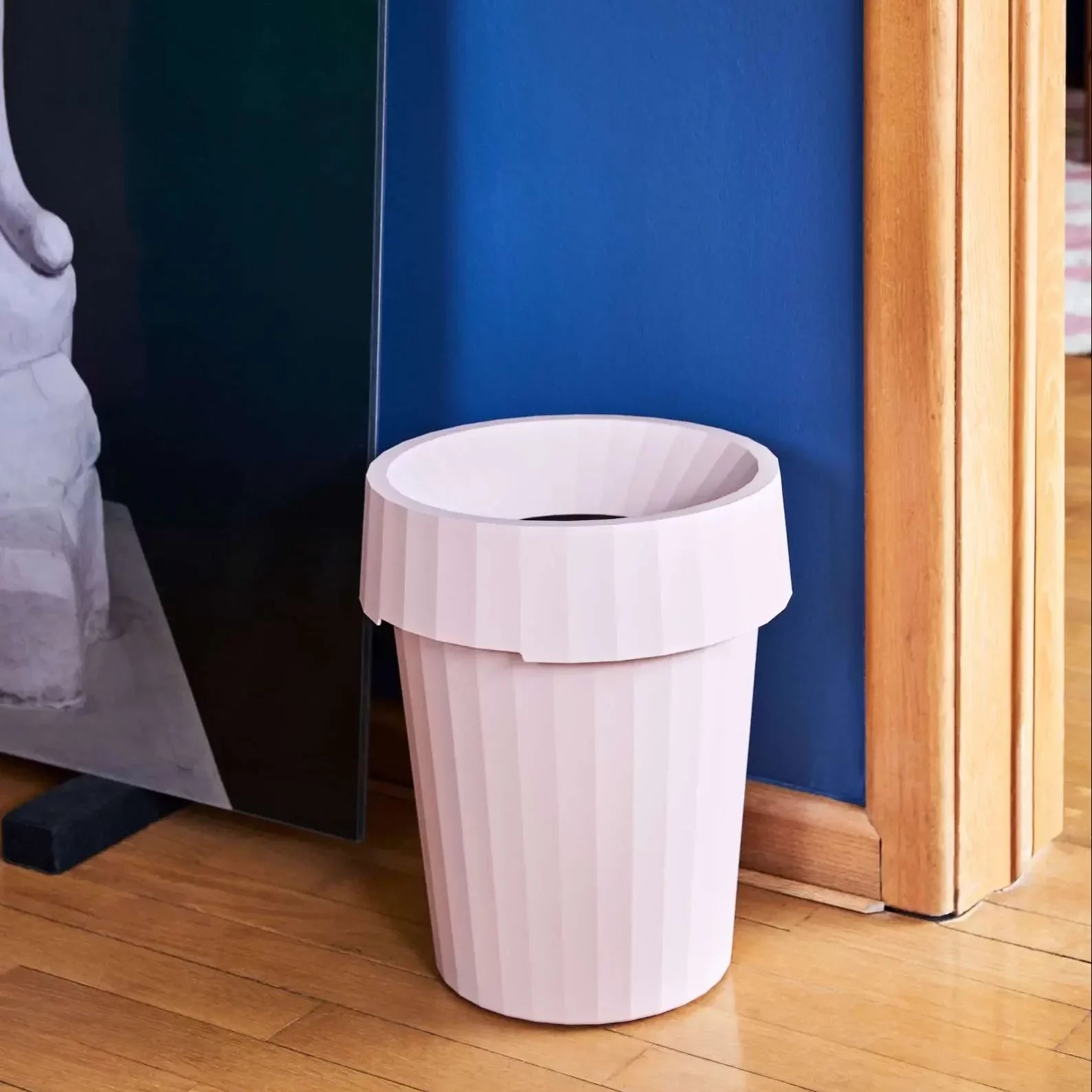 A light pink HAY Shade Bin by HAY, crafted from durable polypropylene with a ribbed design and wide rim, sits on a wooden floor against a blue wall near a door frame.