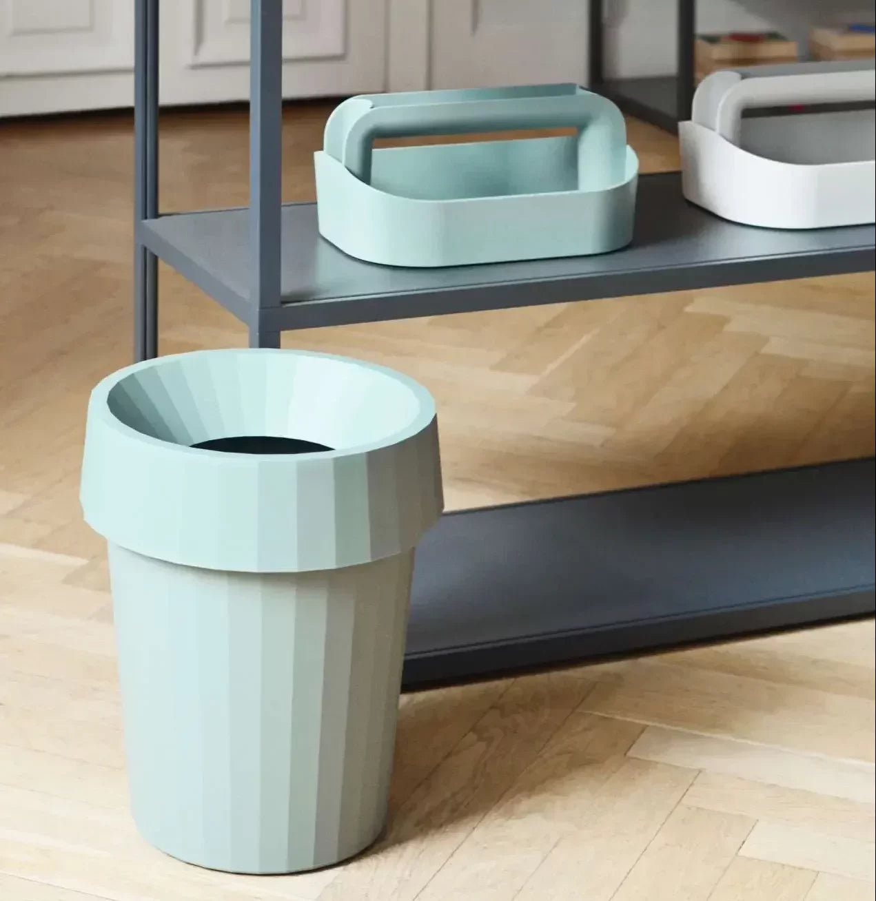 A HAY Shade Bin in light green by HAY, made from durable polypropylene in muted tones, rests on a wooden floor beside a dark grey shelving unit with two stackable baskets—one green, one white.