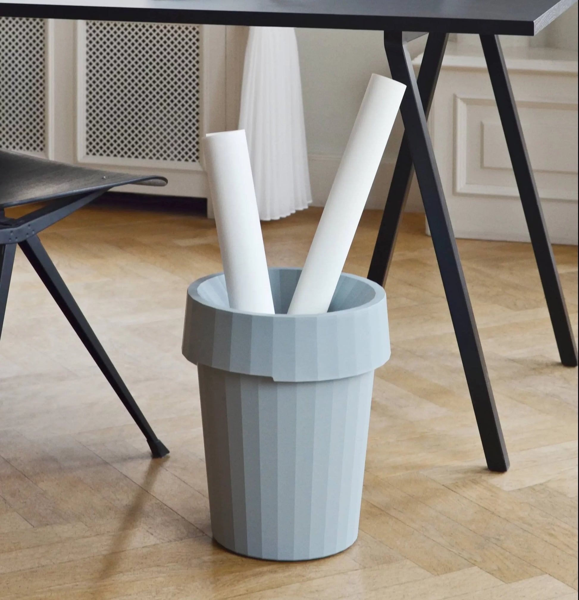 The HAY Shade Bin by HAY, made from durable polypropylene in light gray, sits on a wooden floor under a black table and holds two rolled-up white papers in a modern minimalistic room.