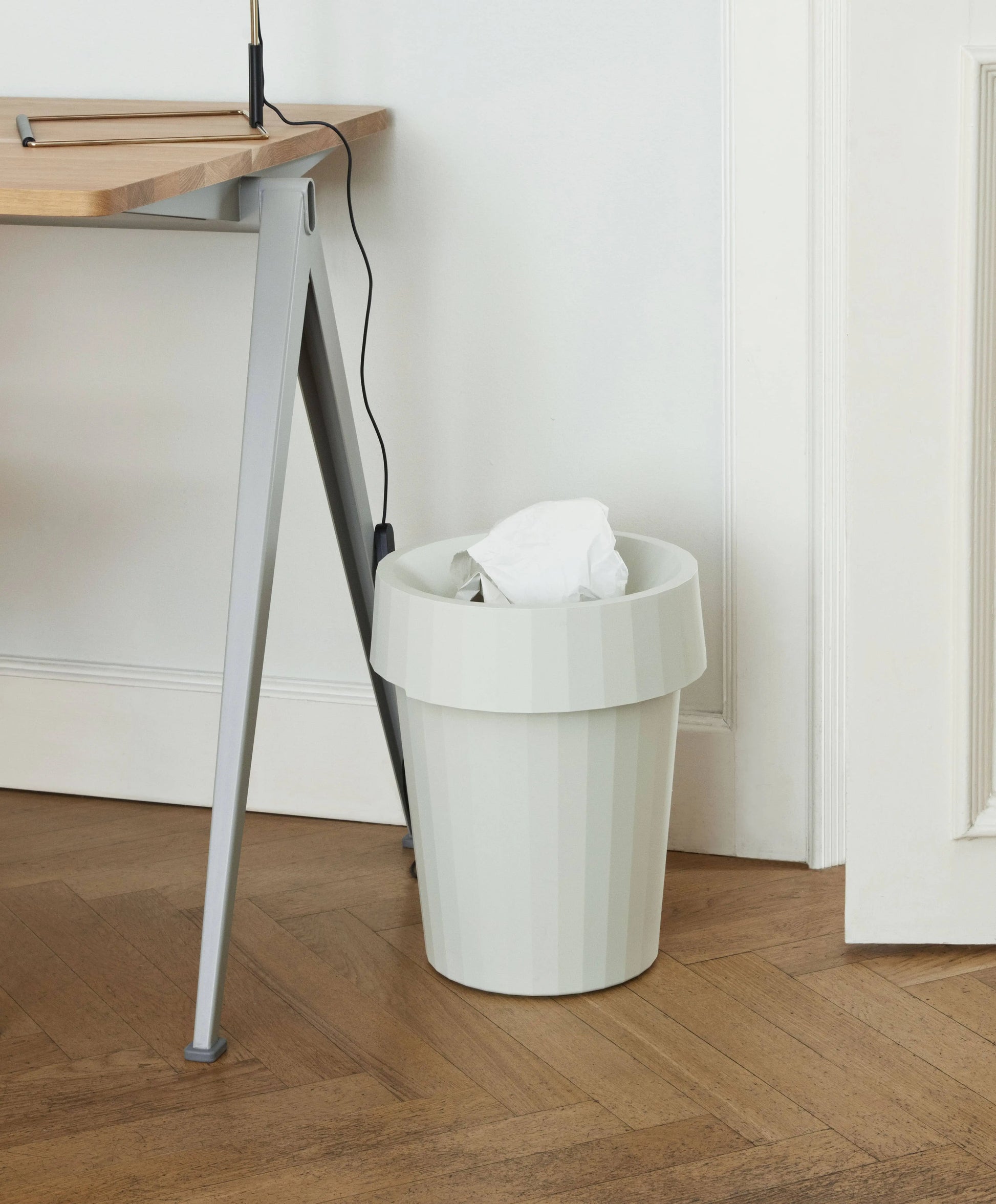 A HAY Shade Bin wastebasket by HAY, made of durable polypropylene in muted white, holds a crumpled paper on a wooden floor near a light desk leg and a wall with a hanging power cord.