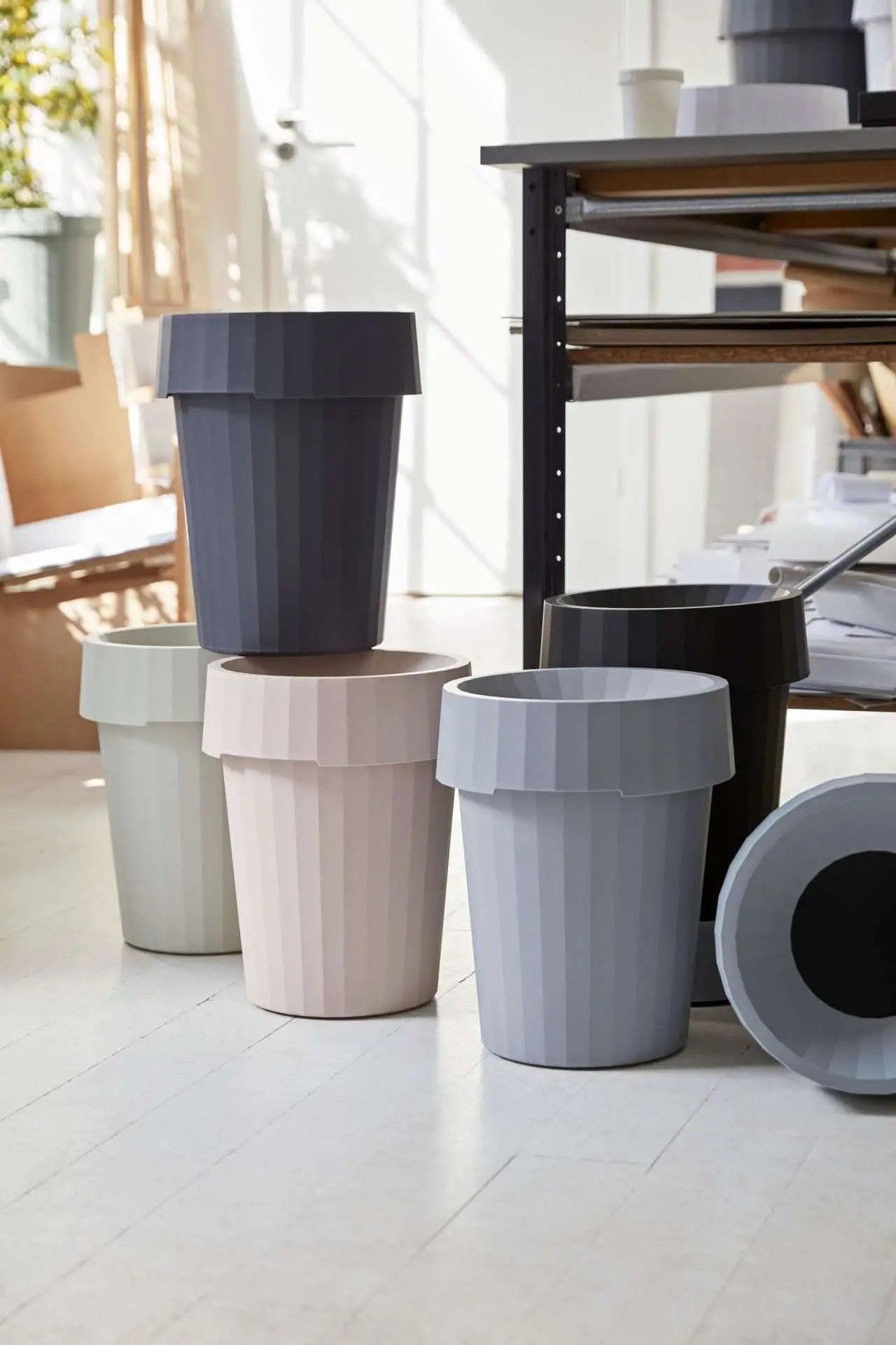 Five HAY Shade Bins by HAY in gray, beige, black, and white durable polypropylene are arranged on a light floor in a bright modern room with shelves and sunlight.