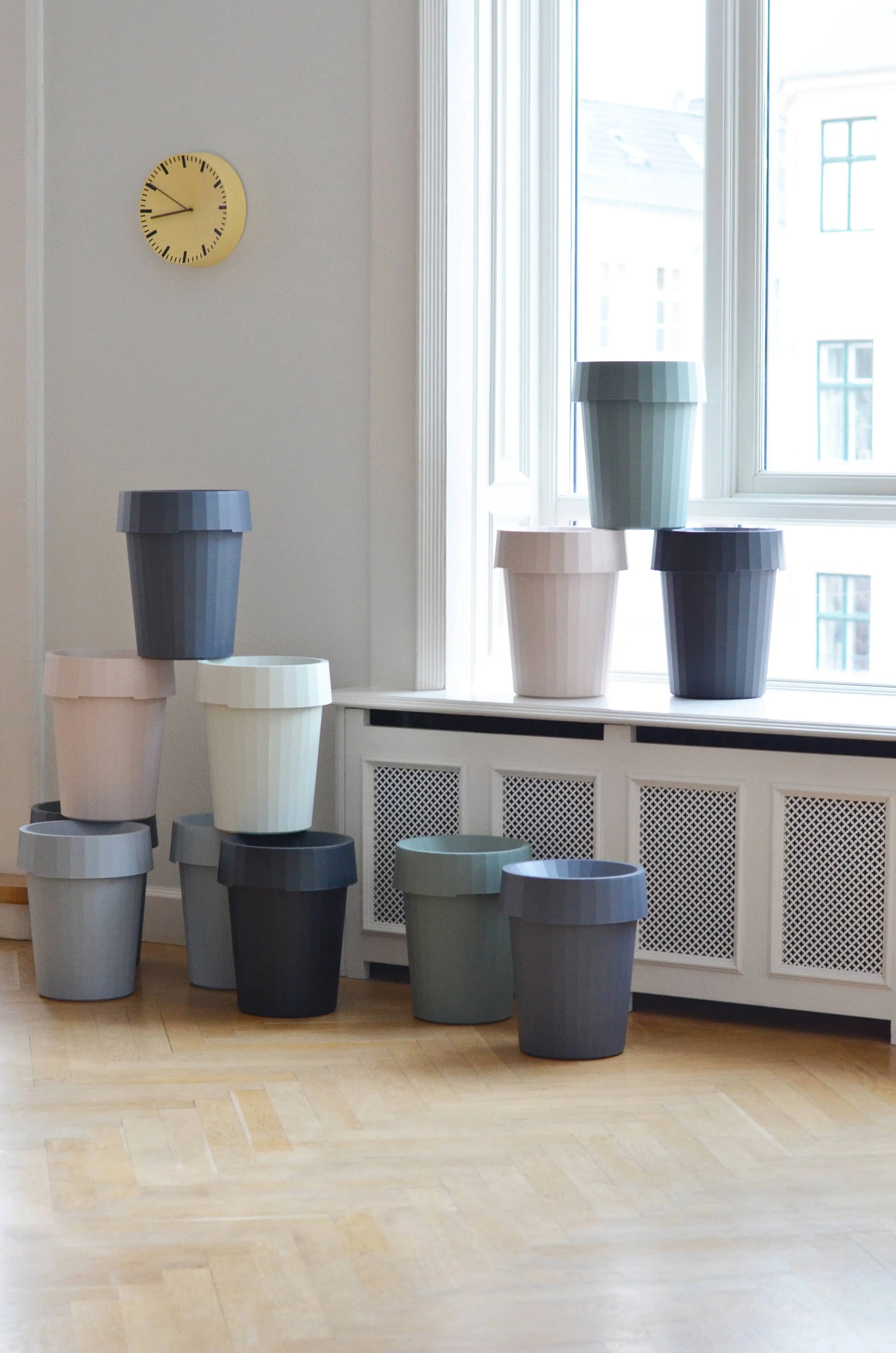 A stack of HAY Shade Bin containers in muted colors sits by a large window on a light wood floor, with a yellow clock above on a white wall. Made from durable polypropylene by HAY.
