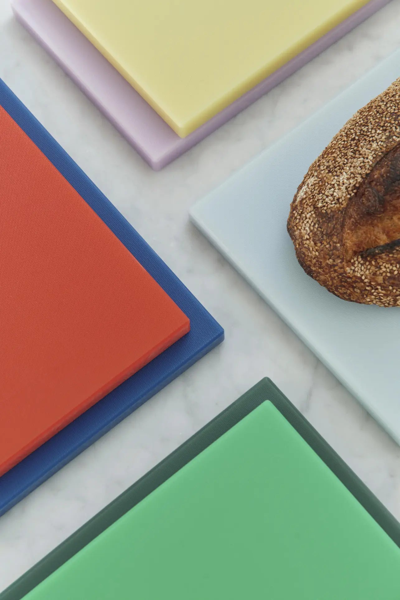 Five colorful HAY Slice Chopping Board L by HAY—in yellow, purple, light blue, red, and green—are displayed on a white marble surface. A sesame-seeded loaf rests partly on the light blue board, illustrating cross-contamination prevention.