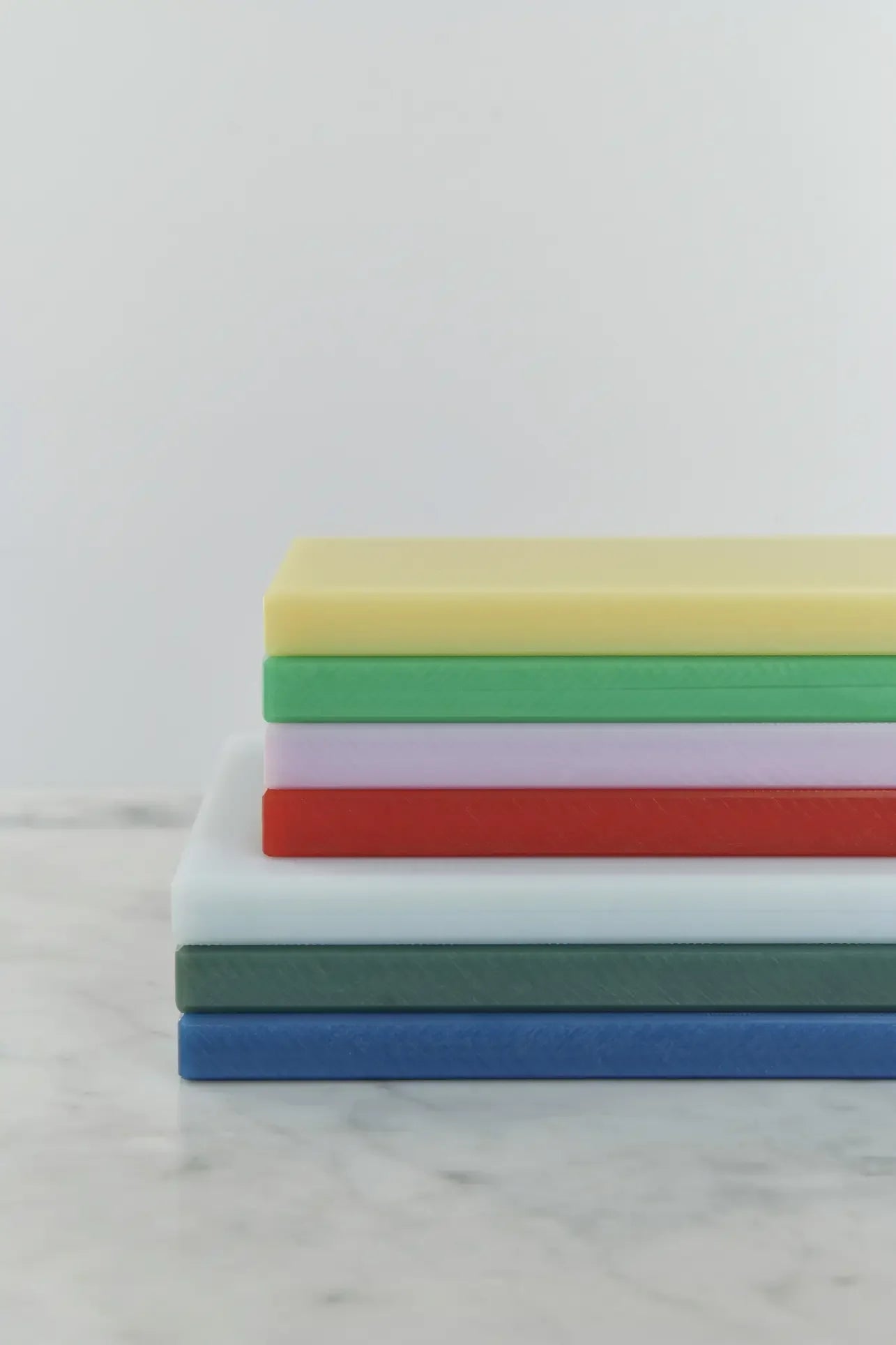 A stack of five HAY Slice Chopping Board L by HAY in yellow, red, green, white, and blue—ideal for preventing cross-contamination—displayed on a light marble surface against a clean white background.