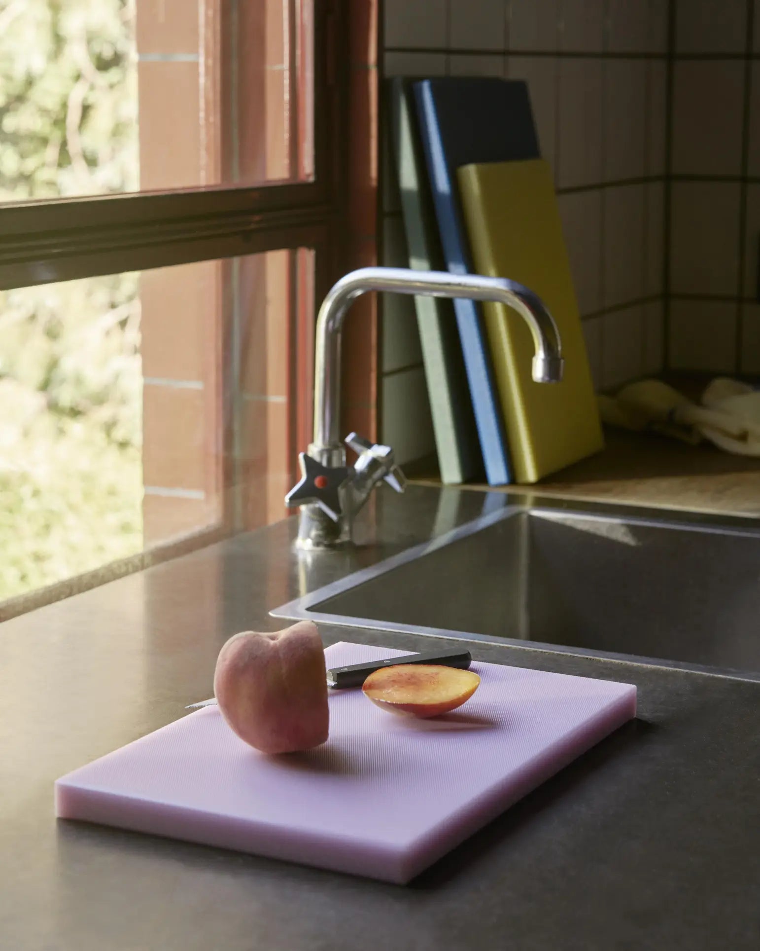 A halved peach rests on a pink HAY Slice Chopping Board L by the sink. Three color-coded kitchen tools from HAY stand nearby to help prevent cross-contamination, illuminated by sunlight through the window.