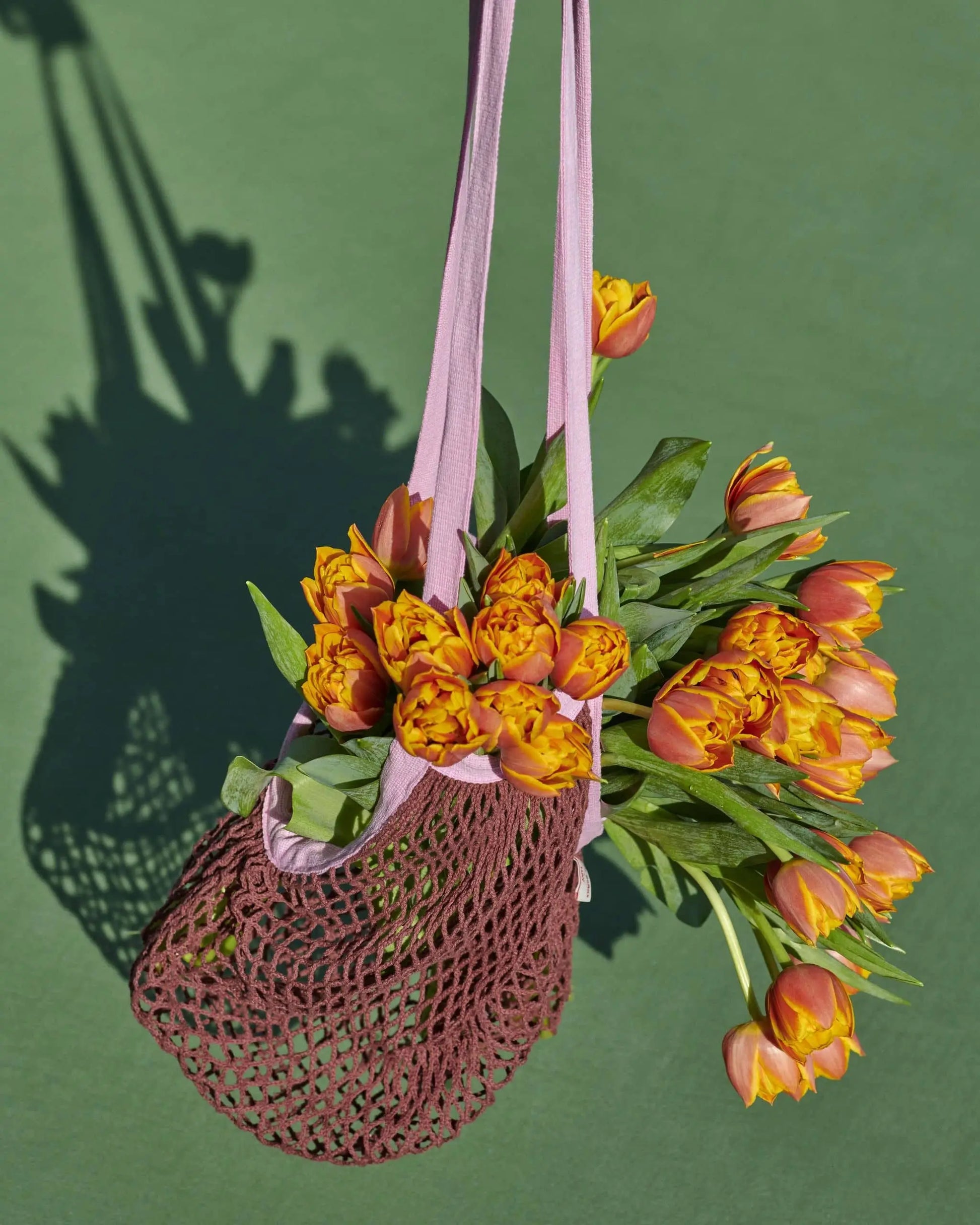 A stylish choice for your groceries, the HAY Sobremesa Market Net Bag by HAY features light pink handles and holds a bouquet of orange tulips with yellow edges, casting a delicate shadow on the green surface.