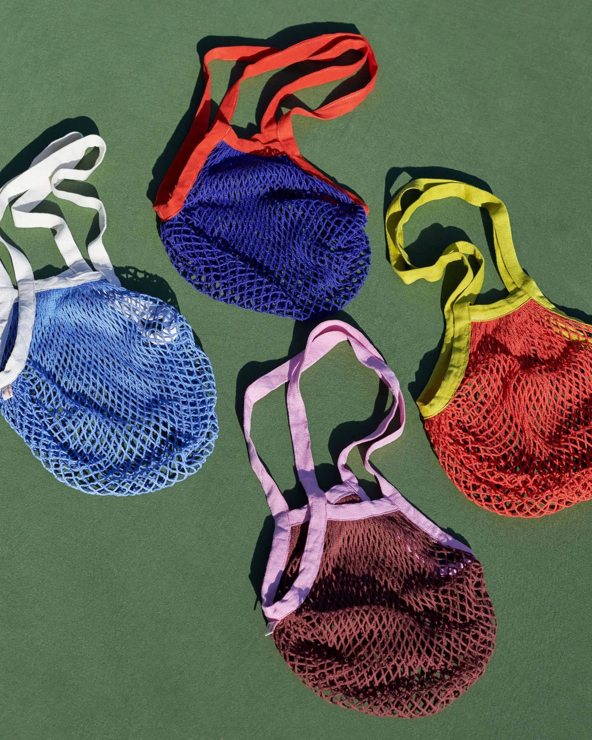 Four HAY Sobremesa Market Net Bags by HAY—in blue, purple, red, and yellow—are laid flat on a green surface. Each stylish netted bag features matching handles, perfect for carrying groceries or daily essentials.