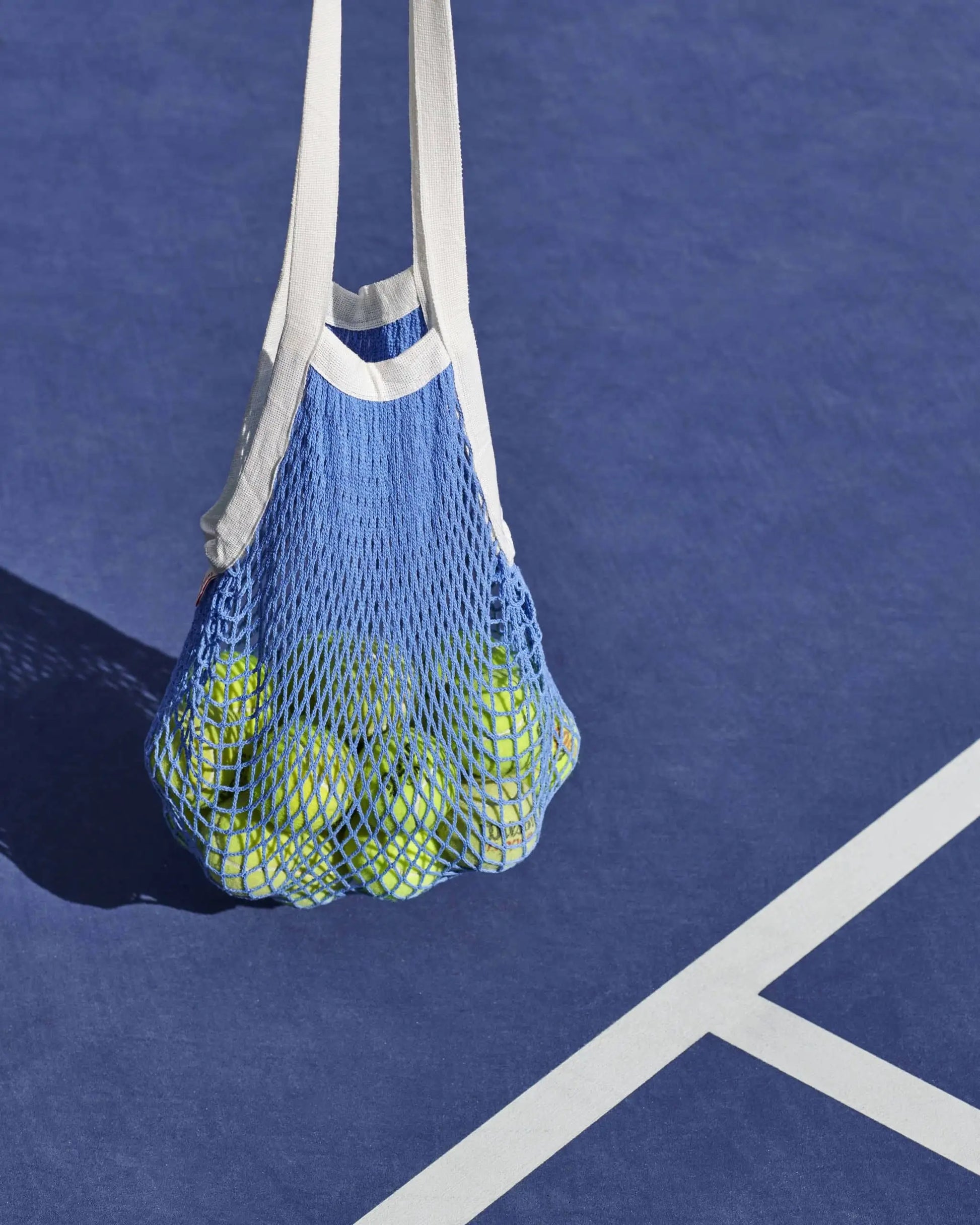 The HAY Sobremesa Market Net Bag by HAY, in blue and white, filled with tennis balls, sits stylishly on a blue tennis court near crisp white boundary lines.