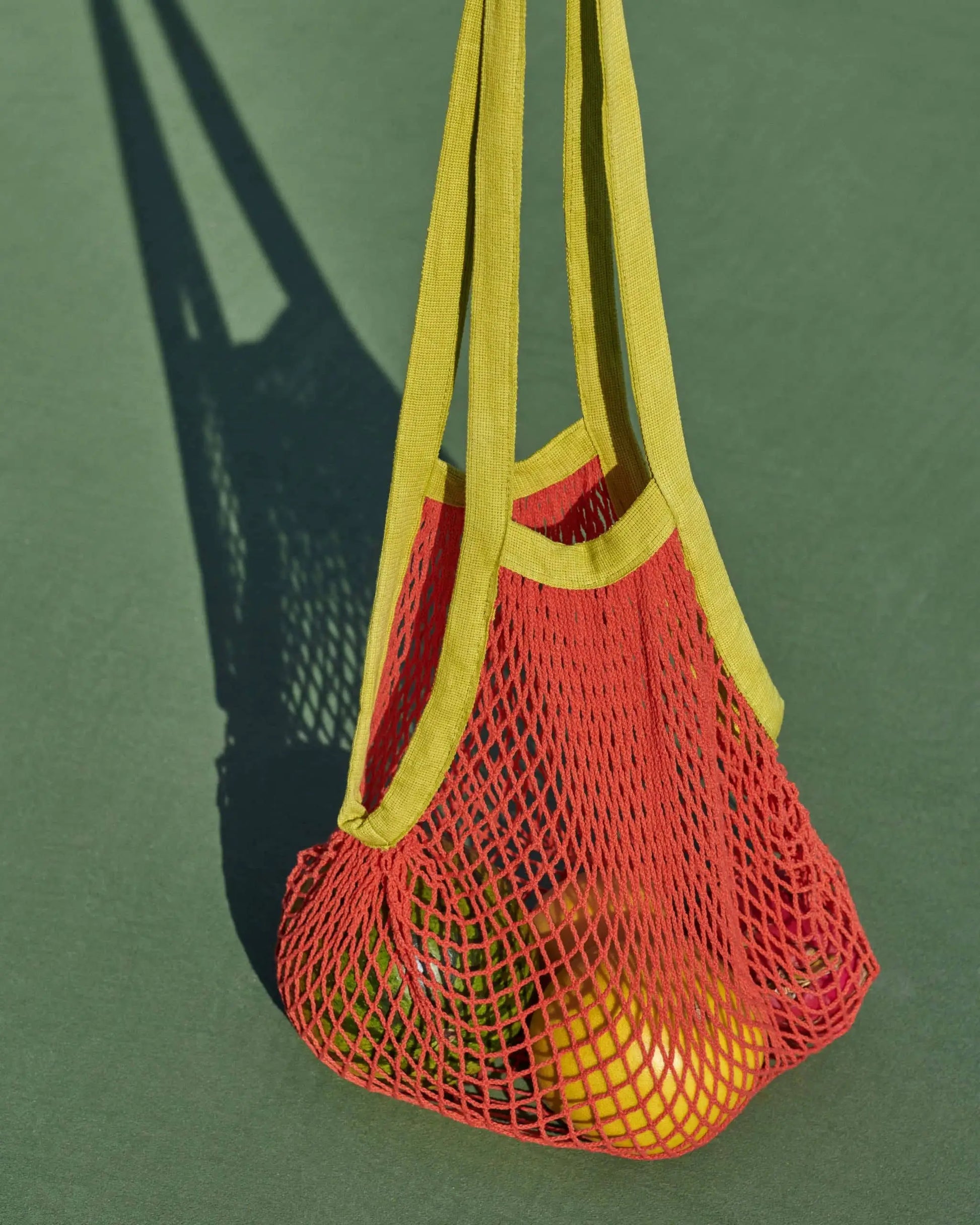 The HAY Sobremesa Market Net Bag by HAY, in red with yellow handles, holds two yellow tennis balls and casts a shadow on a green court—a stylish way to carry more than just groceries.