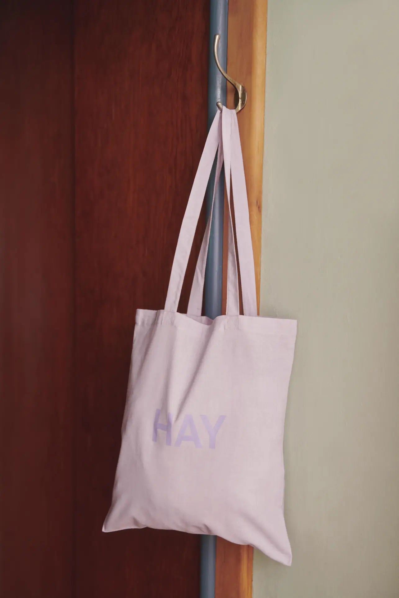 A light pink HAY Tote Bag by HAY, crafted from OEKO-TEX® STANDARD 100 certified organic cotton, hangs on a gold wall hook beside a brown door and a light green wall.