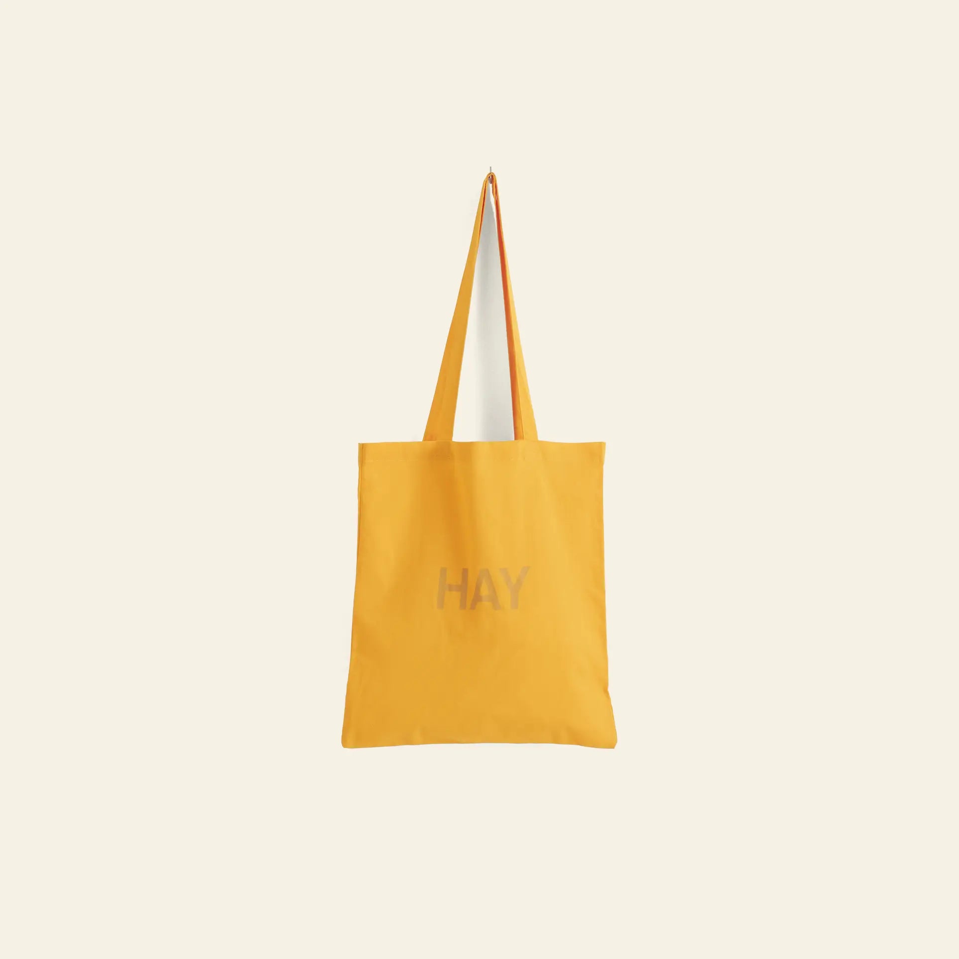 The HAY Tote Bag by HAY is yellow, made from organic cotton, features long handles and a central “HAY” print, shown against a plain light background.