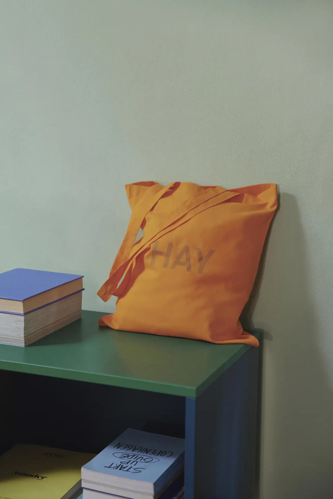 An orange HAY Tote Bag by HAY, made from organic cotton and OEKO-TEX® STANDARD 100 certified, leans against a pale green wall next to a green shelf with stacked books above and partially visible books below.