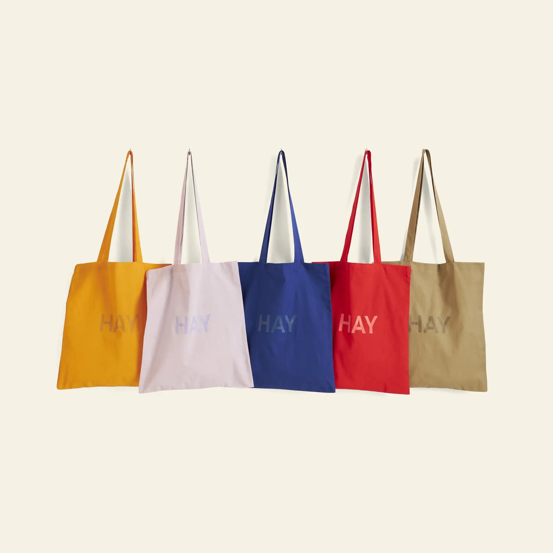 Five HAY Tote Bags by HAY in yellow, light pink, blue, red, and beige are shown in a row. Each organic cotton, OEKO-TEX® STANDARD 100 bag has matching handles and central HAY branding on a plain background.