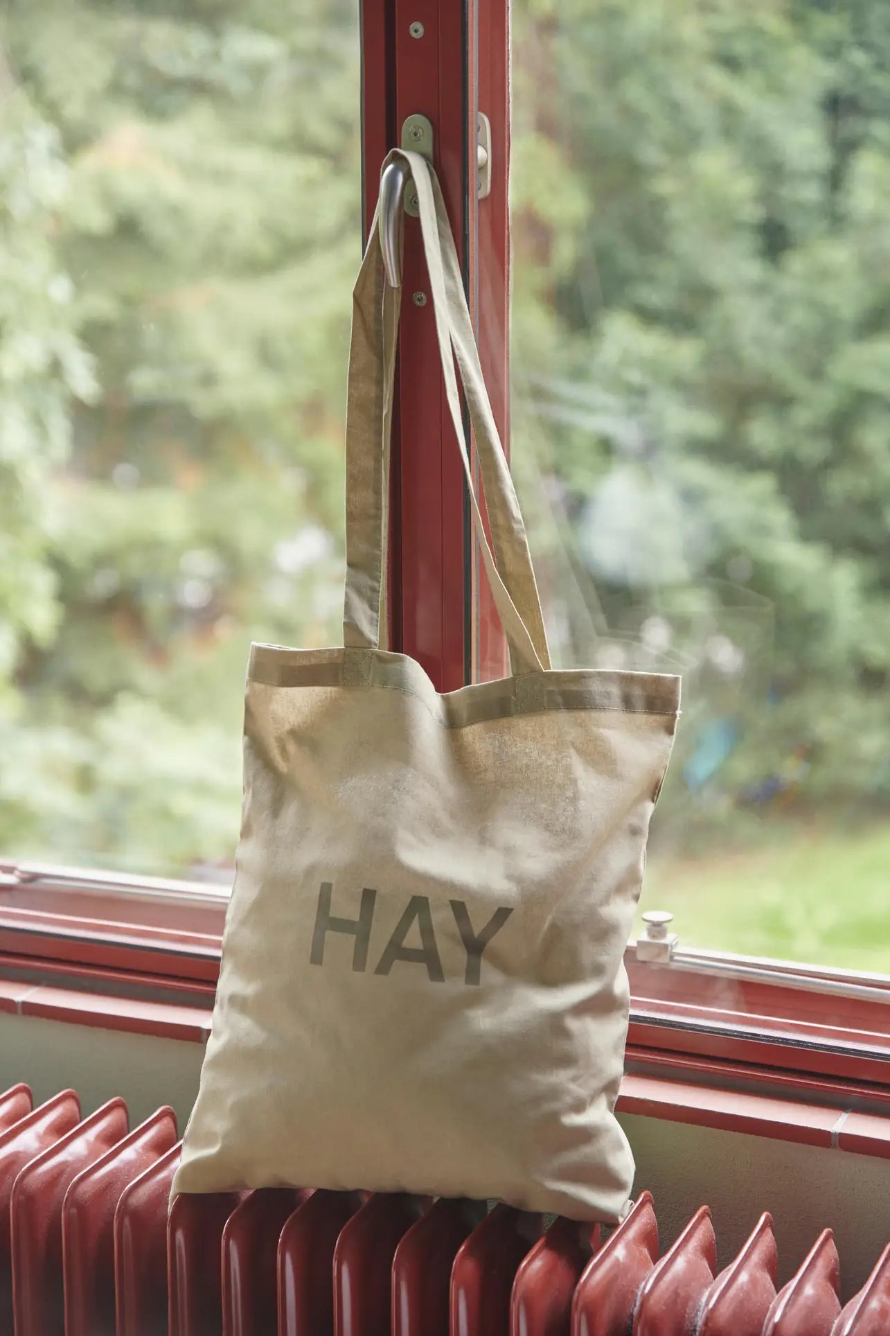 A beige HAY Tote Bag by HAY, crafted from organic cotton and OEKO-TEX® STANDARD 100 certified fabric, hangs from a window latch with lush green trees visible behind the glass and a red radiator beneath.