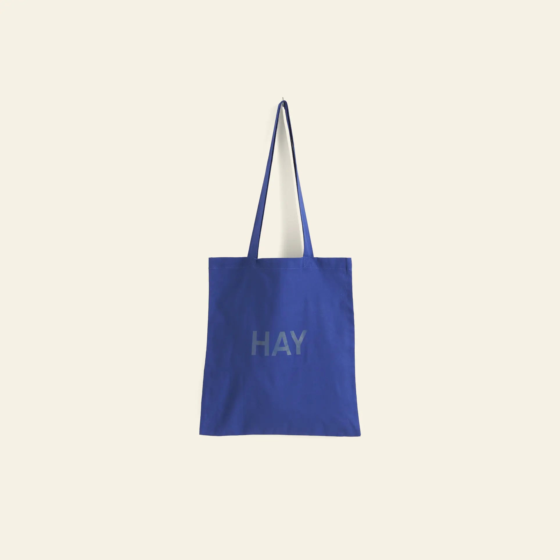 A blue HAY Tote Bag by HAY, made from organic cotton with long handles, hangs on a plain light beige wall. HAY is printed in gray capitals on the front.