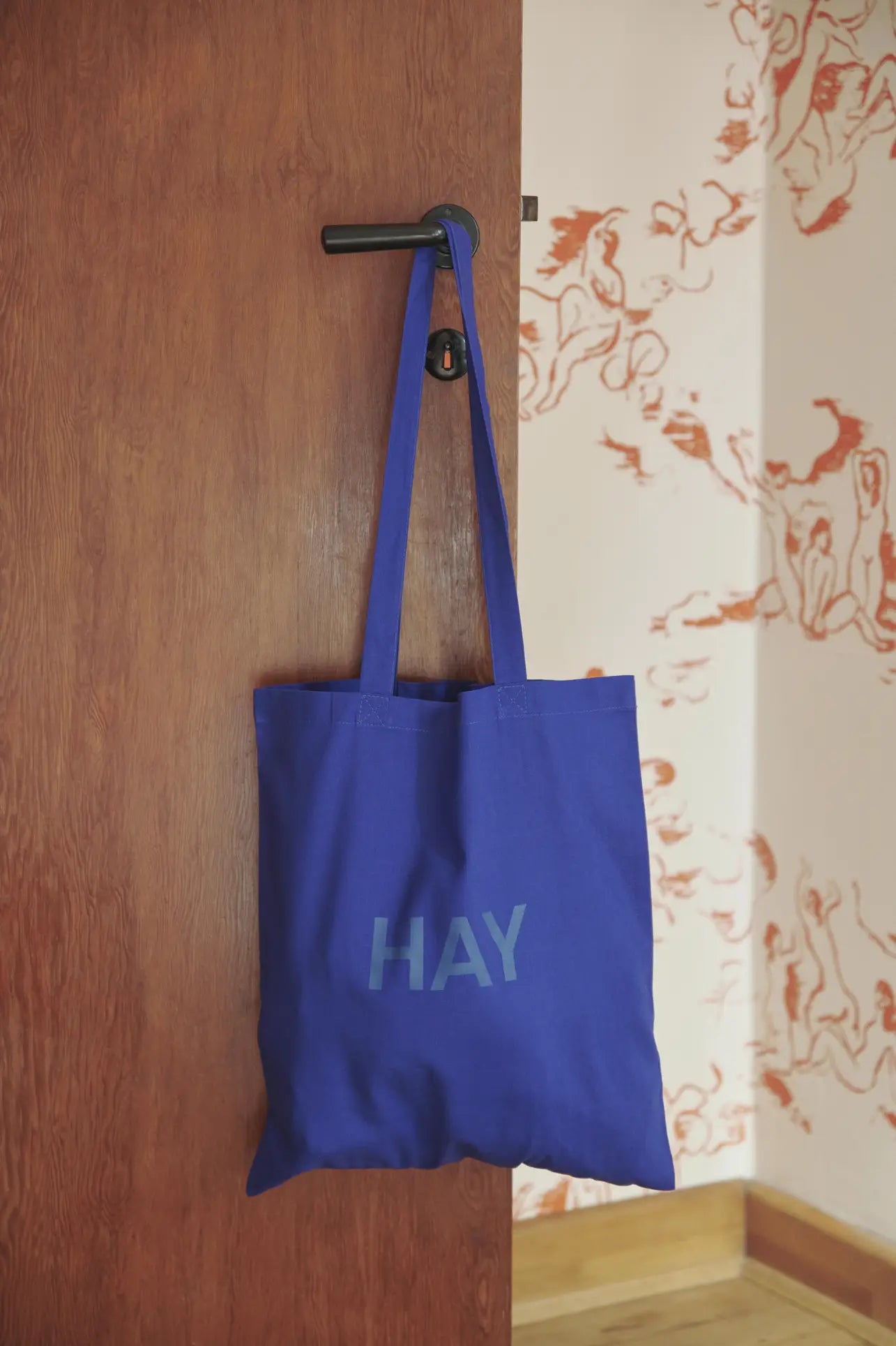 A blue HAY Tote Bag by HAY, crafted from organic cotton and OEKO-TEX® STANDARD 100 certified material, hangs on a black door handle against a brown wooden door and orange abstract wallpaper.