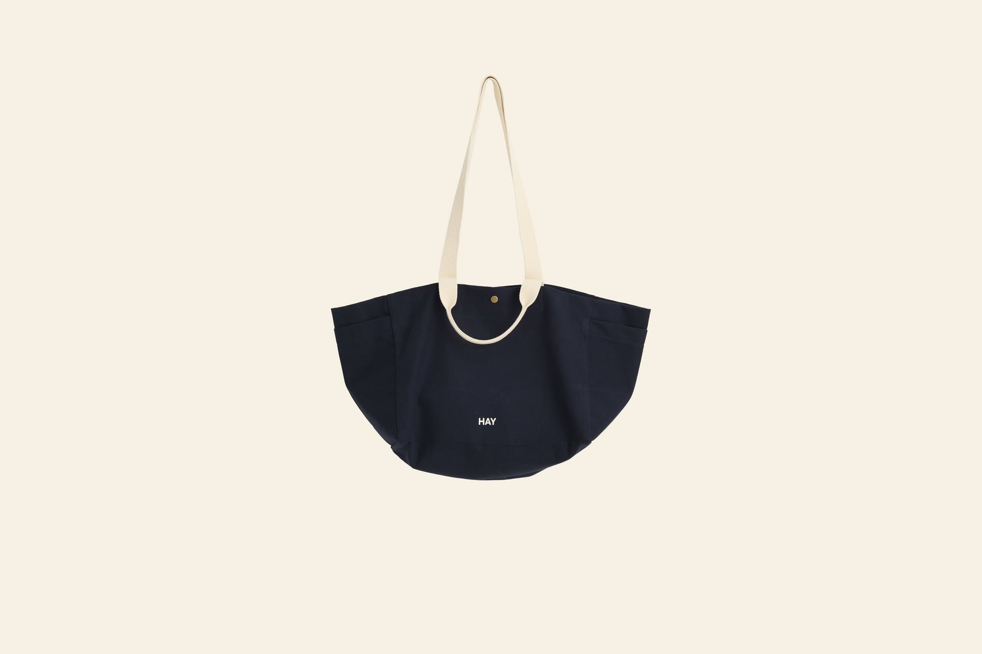 The HAY Weekend Bag Small by HAY is a navy blue organic cotton canvas tote with wide curved sides, cream shoulder straps, a gold snap closure, and MAY in small white letters. Spacious and versatile, its shown against an off-white background.