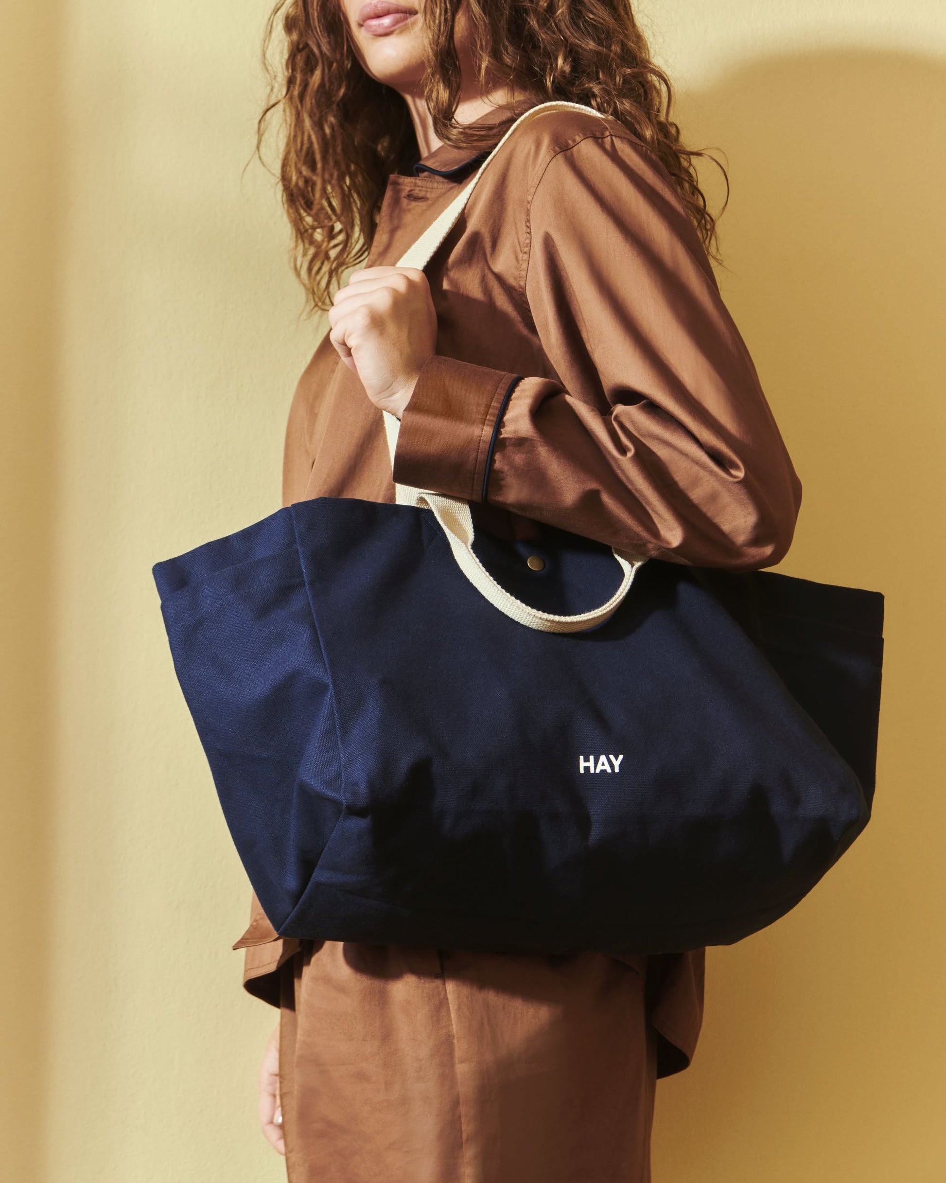 A person in a brown outfit holds the HAY Weekend Bag Small by HAY—a navy blue organic cotton tote with cream handles and HAY print—against a plain light yellow wall, their face partly out of frame.