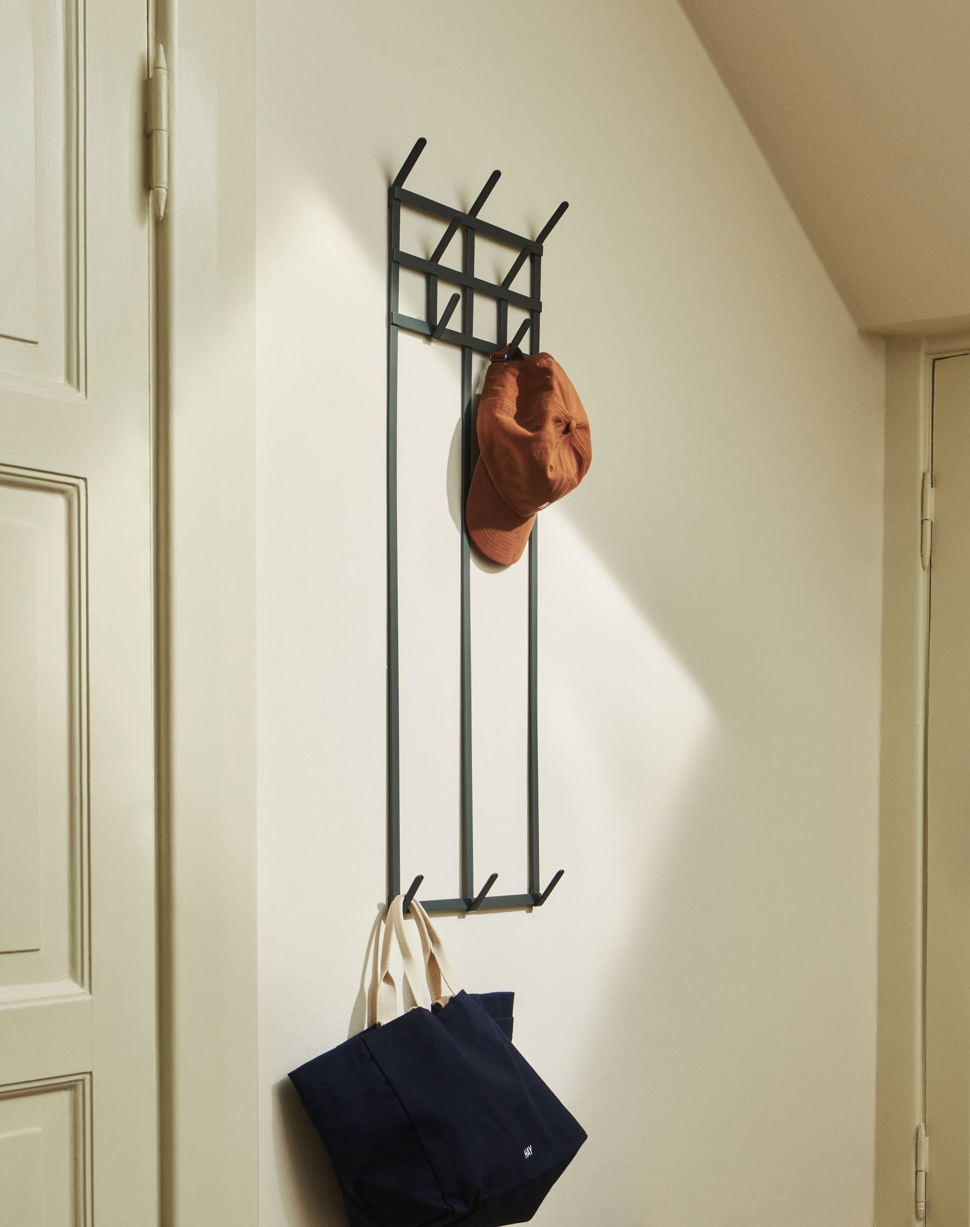 A metal wall-mounted coat rack holds a brown cap and the HAY Weekend Bag Small by HAY, which has a spacious interior, in a bright entryway with white walls and doors.