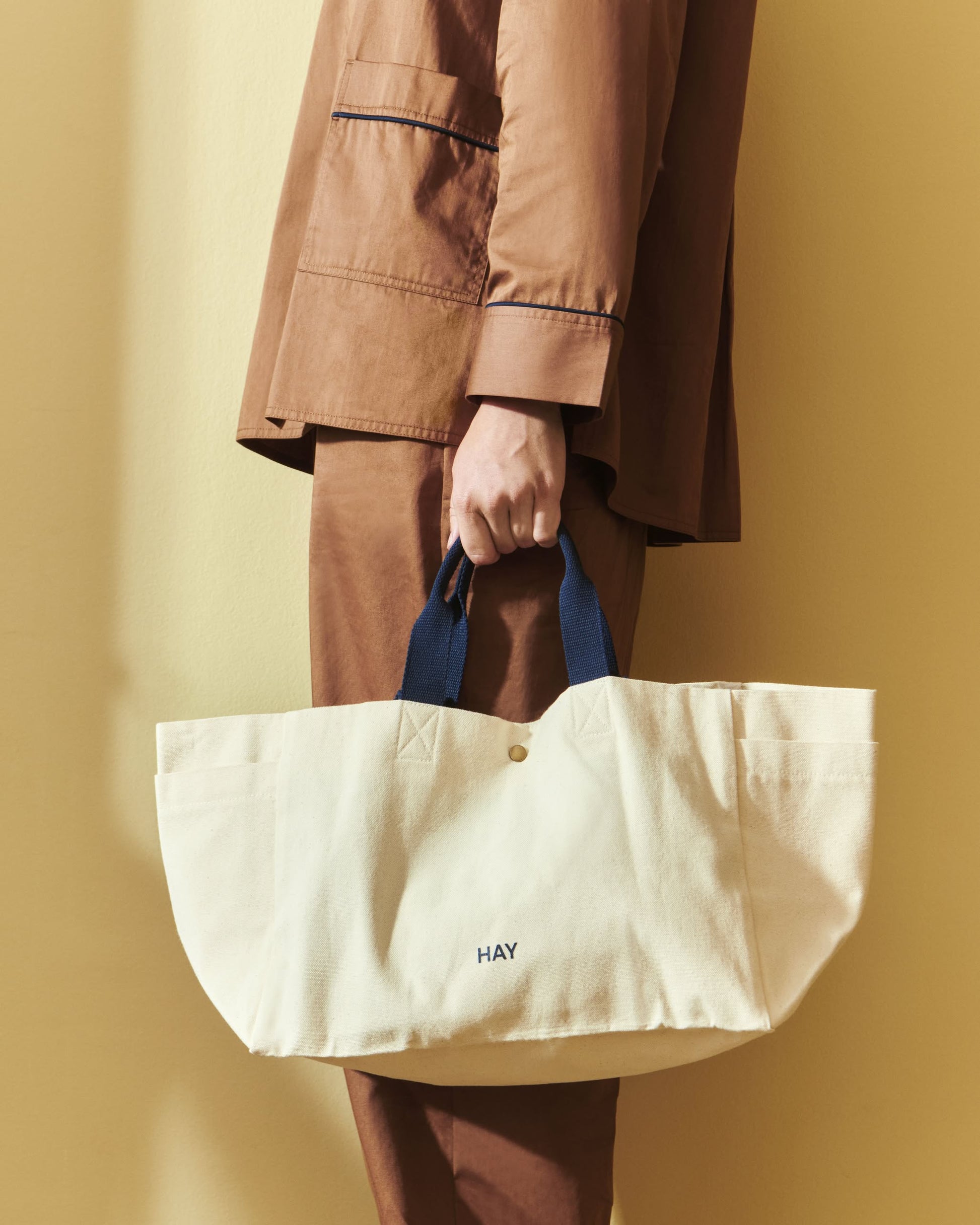 A person in brown stands by a beige wall, holding the HAY Weekend Bag Small by HAY—a cream-colored organic cotton canvas tote with dark handles and “HAY” printed on it, perfect for any outing.