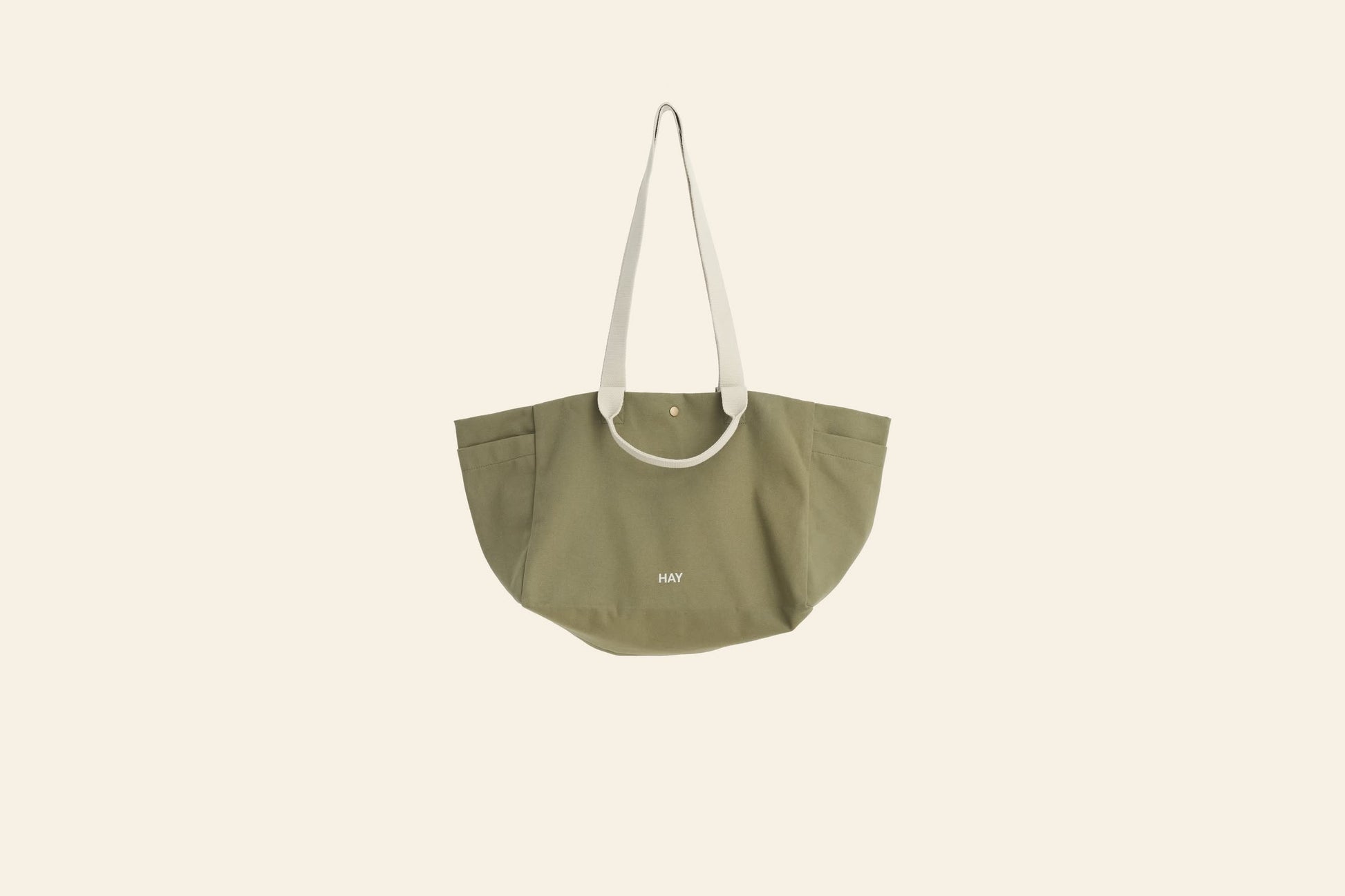 The HAY Weekend Bag Small by HAY is a khaki green canvas tote made from organic cotton, featuring wide off-white shoulder straps and HAY printed in small white letters on the front, shown against a plain cream background.