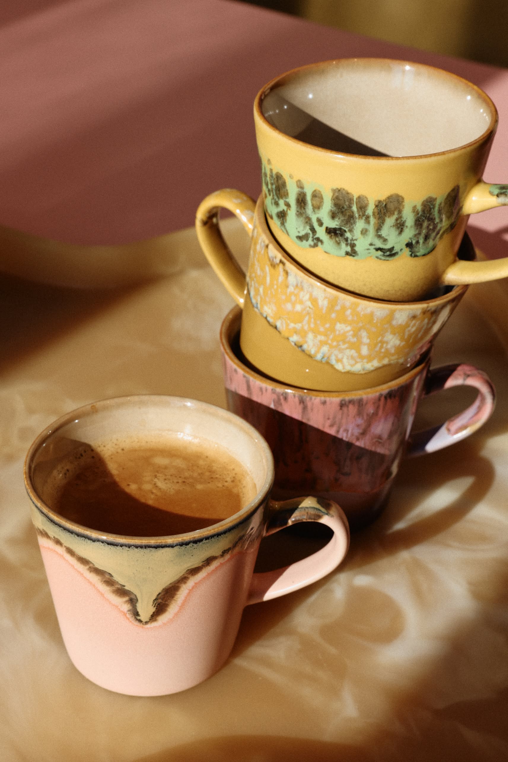 Four HKliving 70s Ceramics: Americano Mugs are stacked on a beige surface, each with a unique design from HKliving. In front, a pink mug filled with coffee stands out as warm sunlight casts soft shadows over the scene.