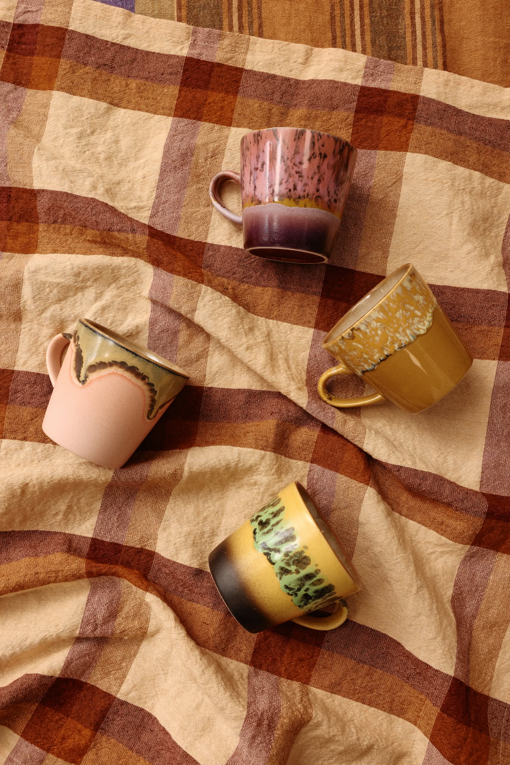 The HKliving 70s Ceramics: Americano Mugs Set of 4 features retro coffee mugs in pink, yellow, green, and earthy glazes. Each uniquely patterned mug is arranged on brown plaid fabric, showcasing the vintage charm of HKliving ceramics.
