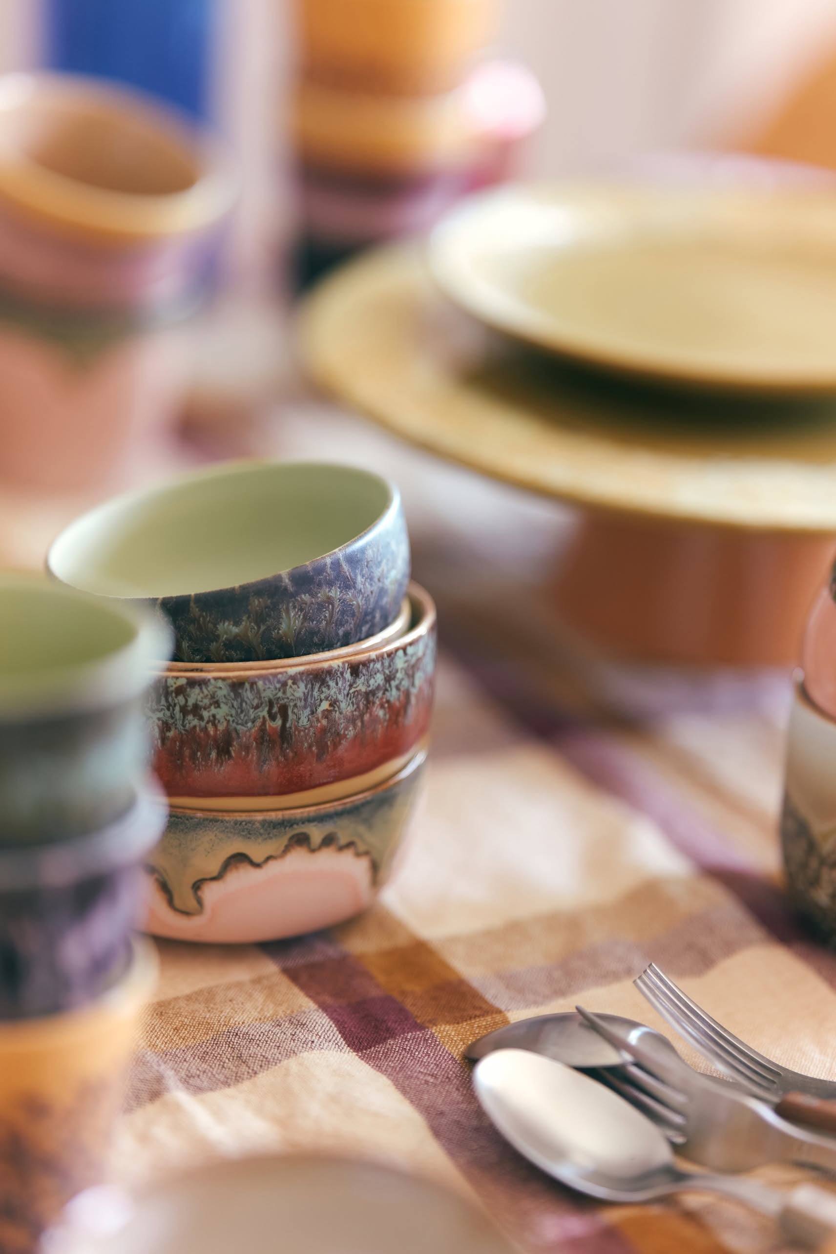 A close-up of HKliving 70s Ceramics: Dessert Bowls, Set of 4, featuring colorful retro designs stacked on a plaid tablecloth, with plates, spoons, and forks in the background to create a cozy table setting.