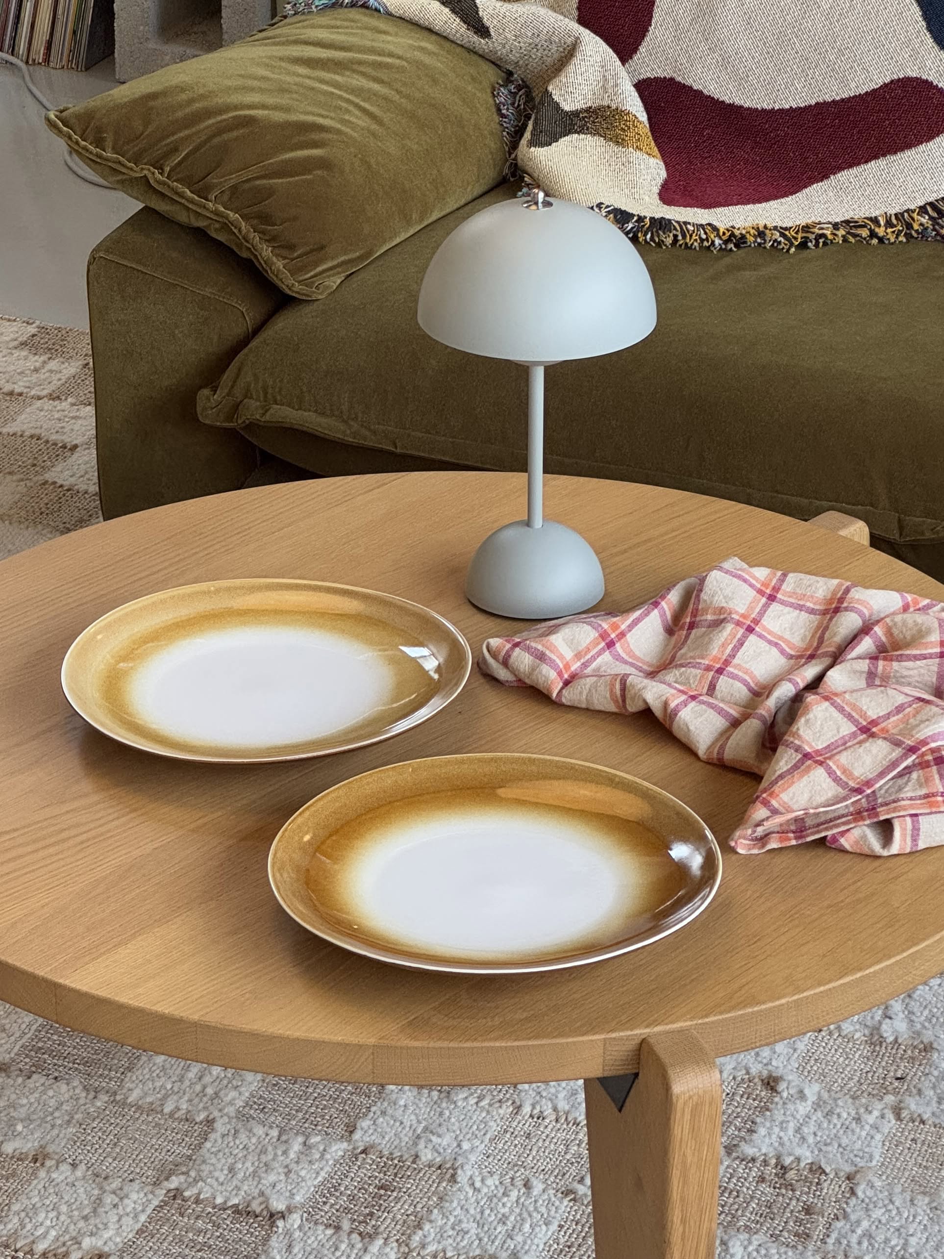 Two HKLIVING 70s Ceramics: Dinner Plates, Set of 2 - Supernova, with hand-glazed finishes, rest on a round wooden table beside a folded pink plaid cloth and modern gray lamp, with a green couch and patterned blanket in the background.