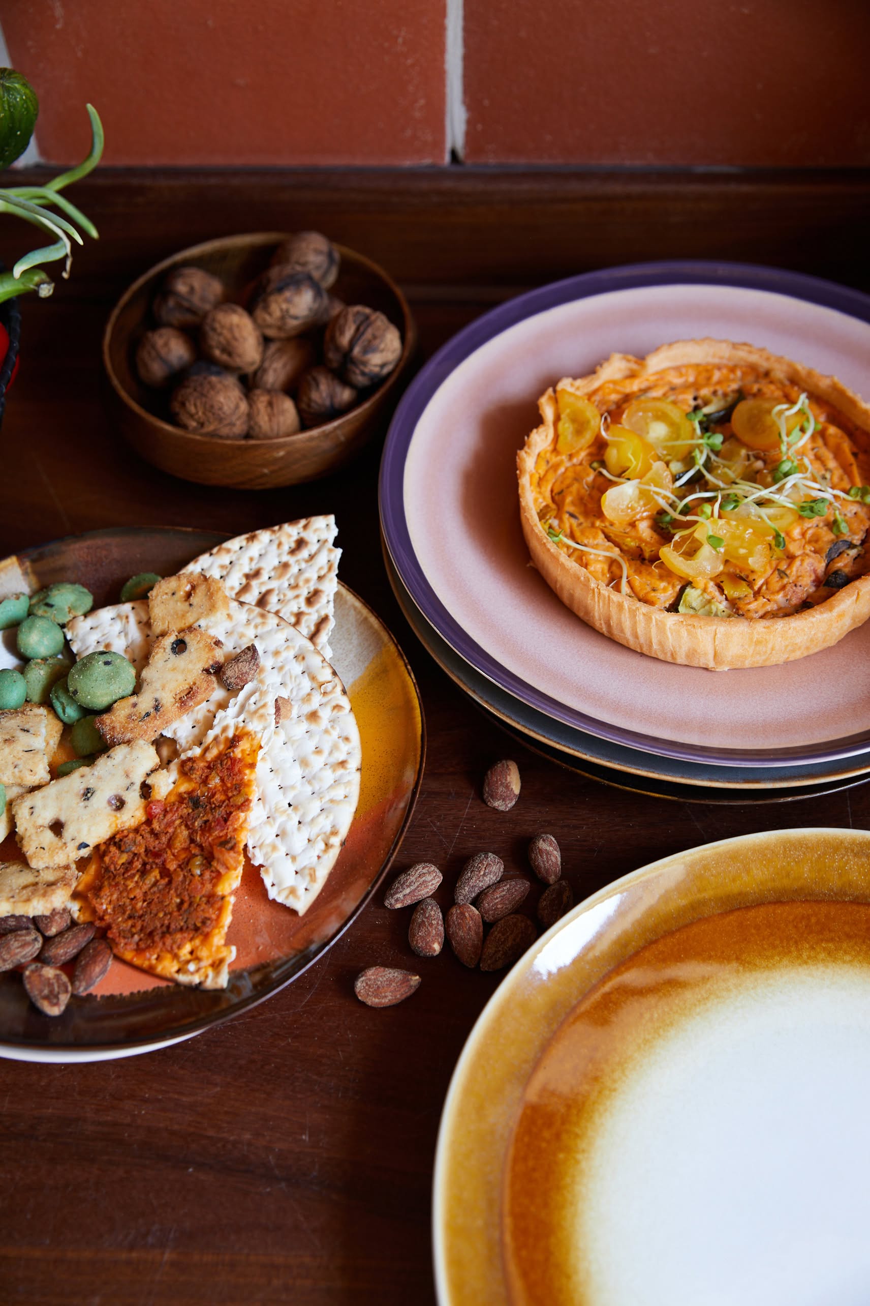 A table set with HKLIVING 70s Ceramics: Dinner Plates, Set of 2 - Supernova, topped with assorted crackers, a tart with yellow tomatoes and greens, walnuts in a bowl, and scattered almonds against a brown tile background.