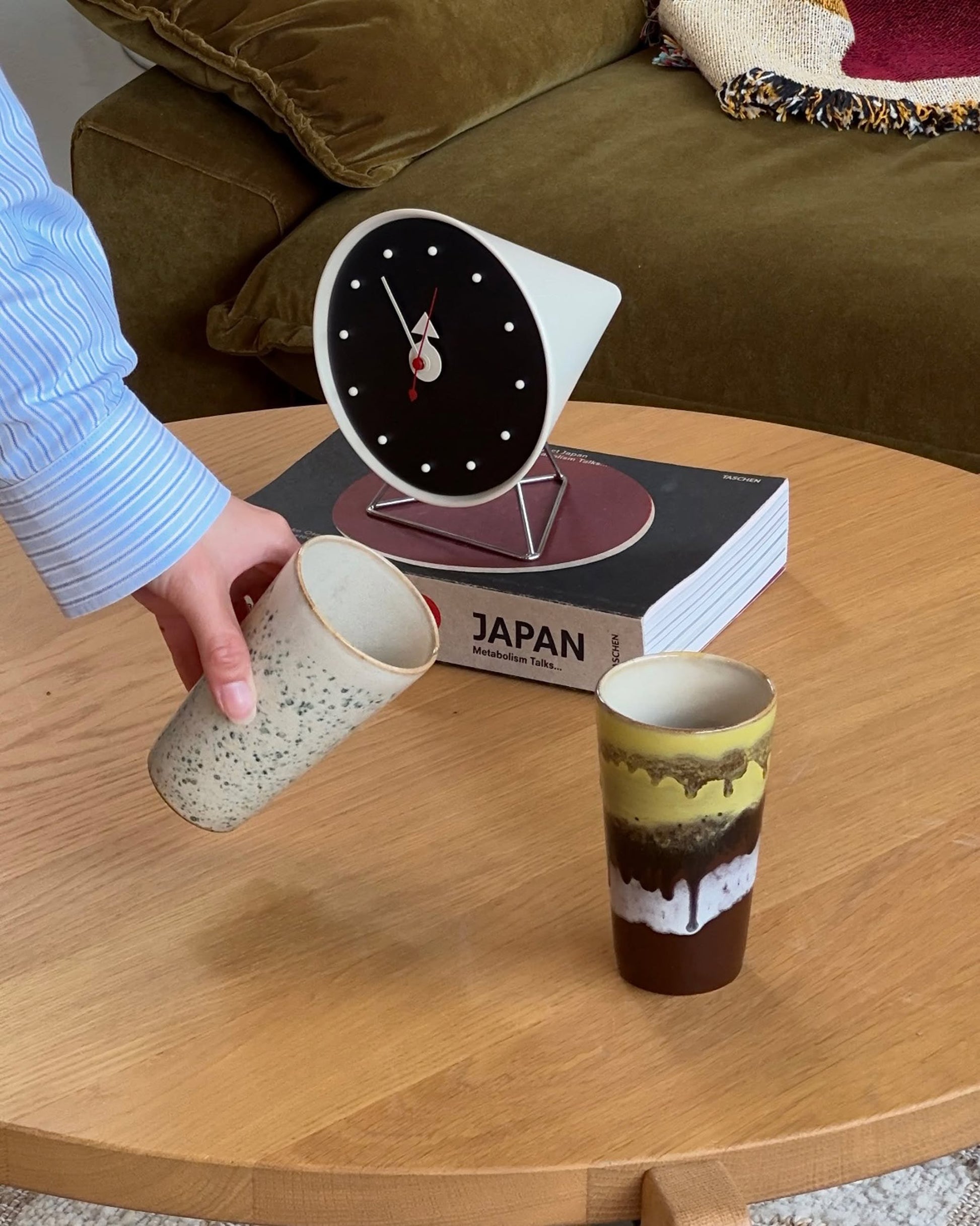 A hand reaches for an HKliving 70s Ceramics: Latte Mug on a wooden table beside its matching mug, with a clock, the book “JAPAN,” and a green couch with a blanket in the background.
