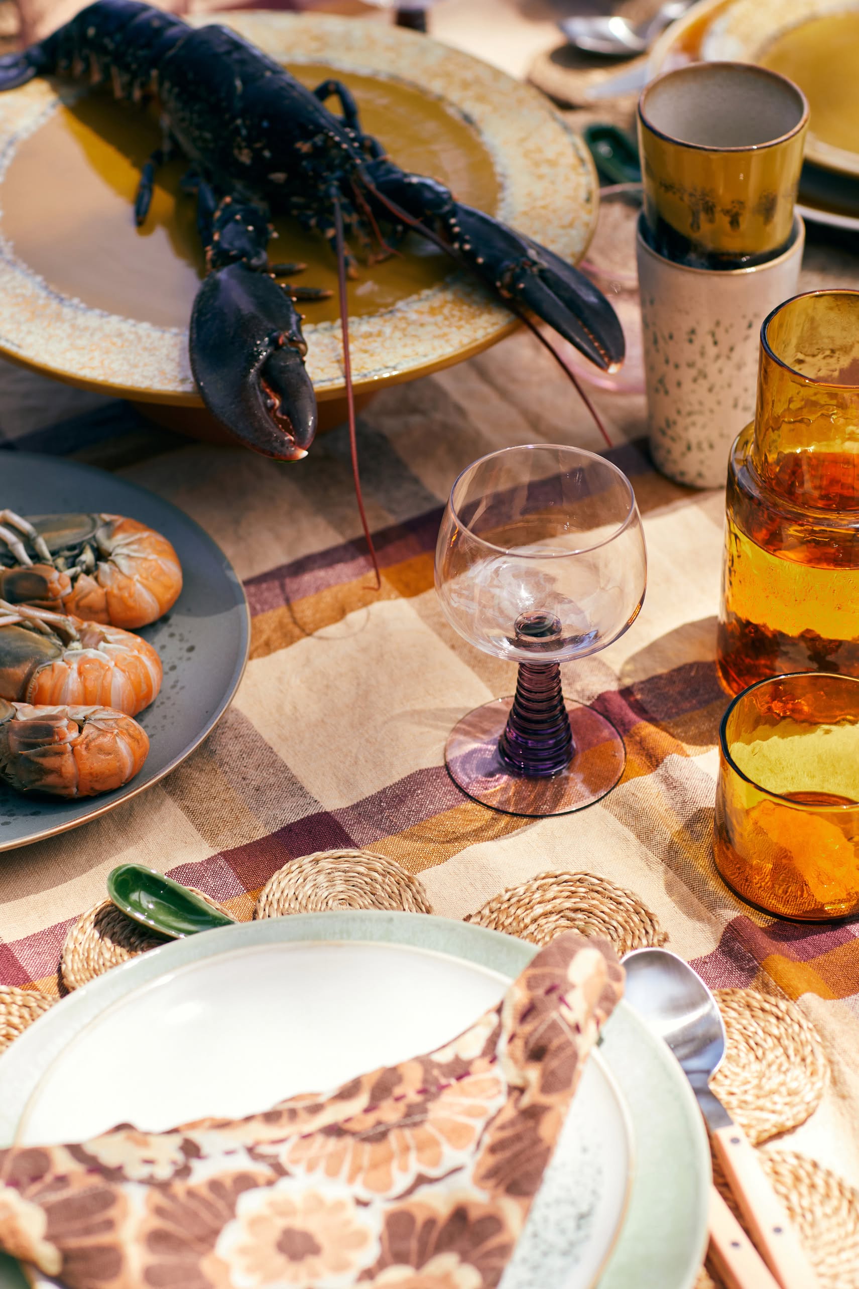 A table set for a meal features a large lobster, prawns, an empty wine glass, amber glasses, and an HKliving 70s Ceramics Latte Mug from the set of 2, all atop woven placemats and a patterned tablecloth.