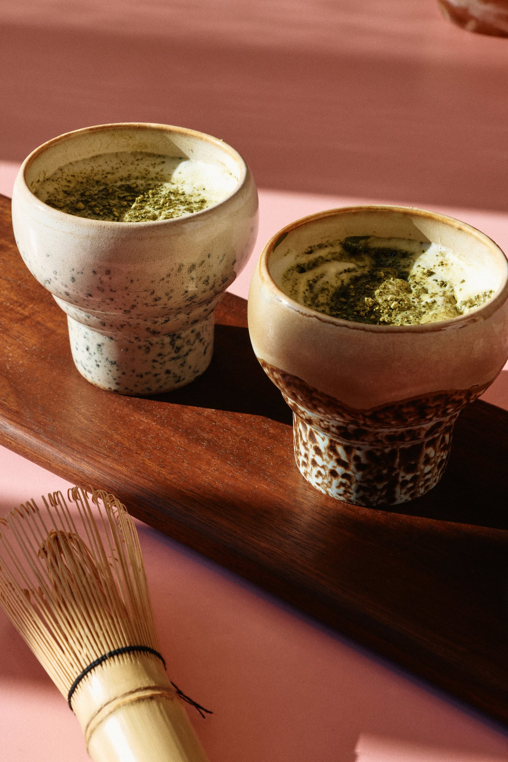 The HKLIVING 70s Ceramics: Lungo Mugs, Set of 2 sit on a wooden tray with frothy matcha, accompanied by a bamboo whisk on a pink surface—ideal for coffee and tea lovers seeking retro-inspired style.