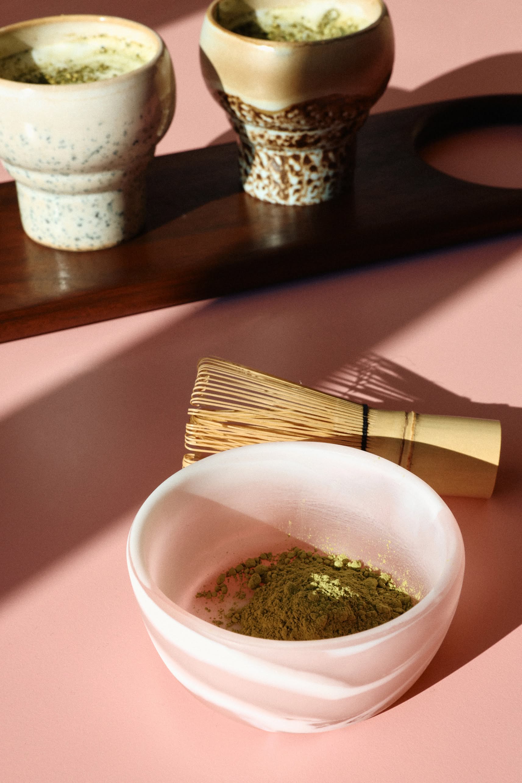 A white bowl with green matcha powder and a bamboo whisk sits on a pink surface. In the background, two cups of matcha tea rest beside HKLIVING 70s Ceramics: Lungo Mugs, Set of 2, as sunlight casts soft shadows across the scene.
