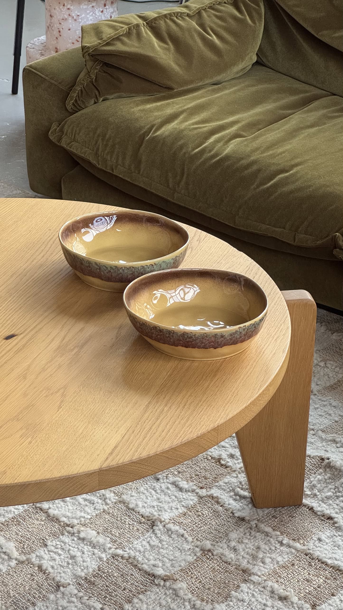 Two HKliving 70s Ceramics Pasta Bowls, Set of 2, with subtle floral designs, sit on a light wood coffee table before an olive-green sofa. A textured beige rug is partially visible on the floor.