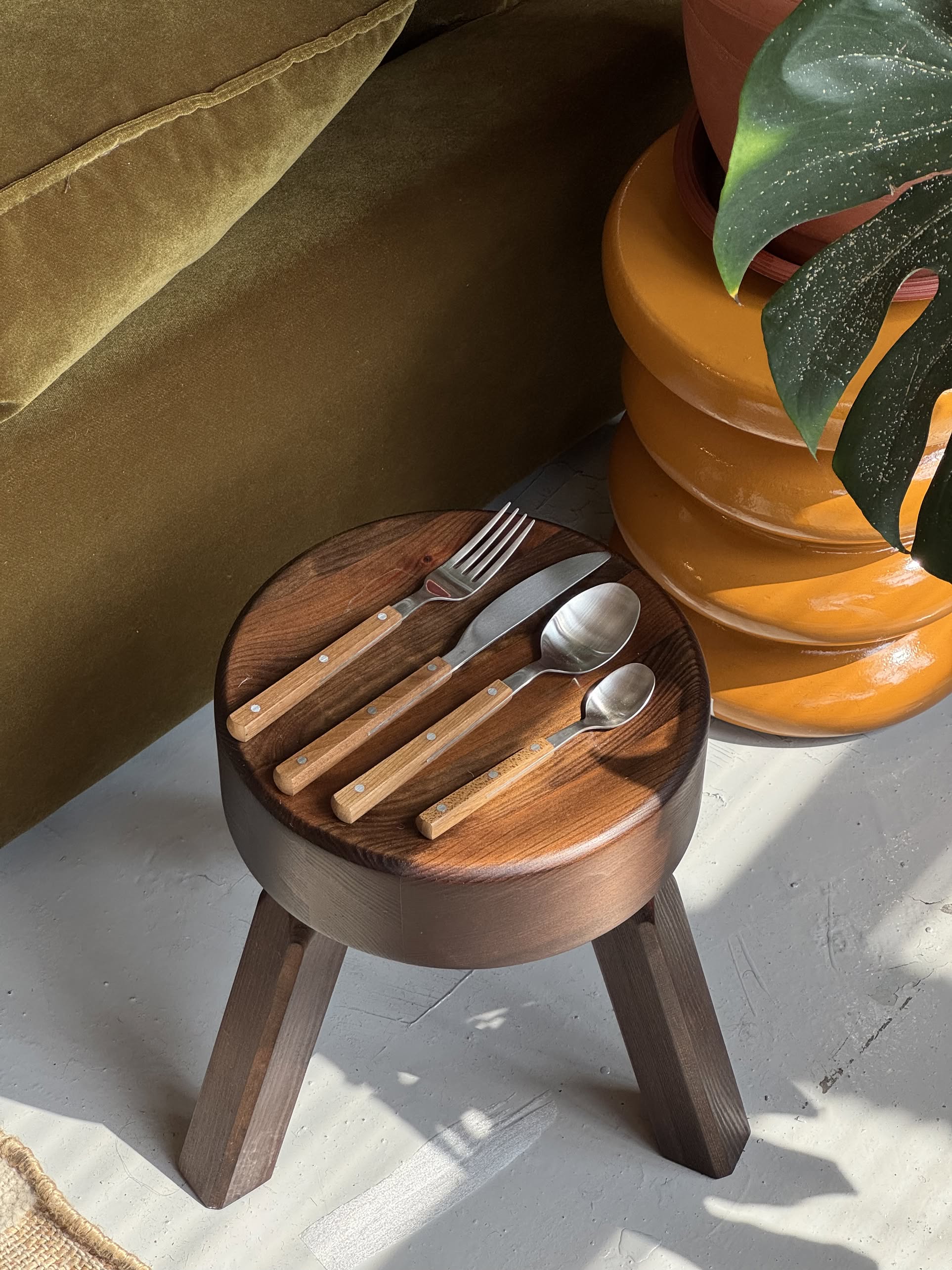 A HKLIVING 70s Cutlery Set of 16 (wood) rests on a wooden stool beside a green velvet couch and a yellow side table with a potted plant, as sunlight casts soft shadows on the floor.