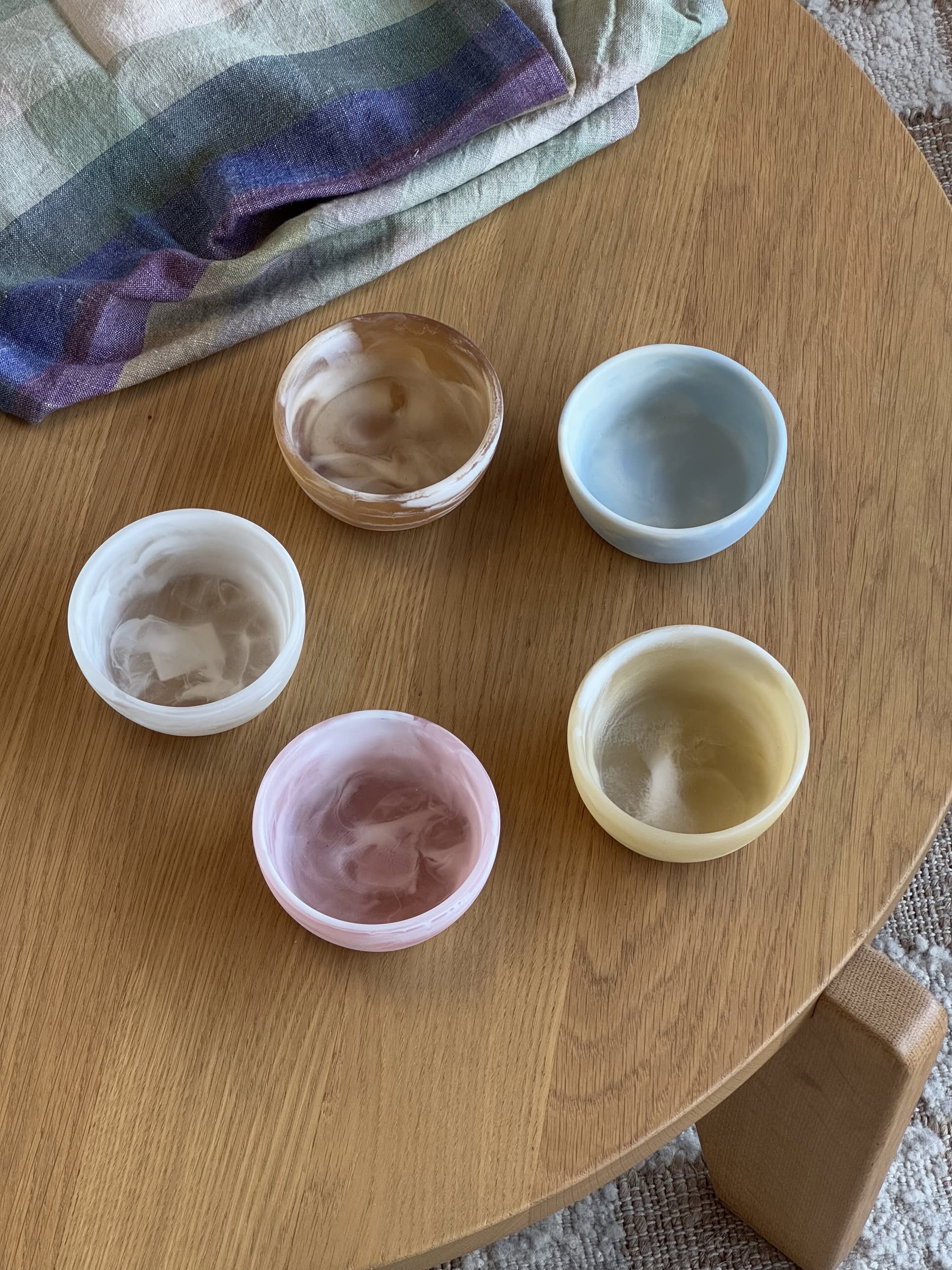 Five HKLIVING Patio Bowls by HKLIVING in pastel marbled resin (brown, blue, white, pink, yellow) are neatly arranged on a wooden table—ideal for outdoor entertaining. A plaid cloth appears in the upper left corner.