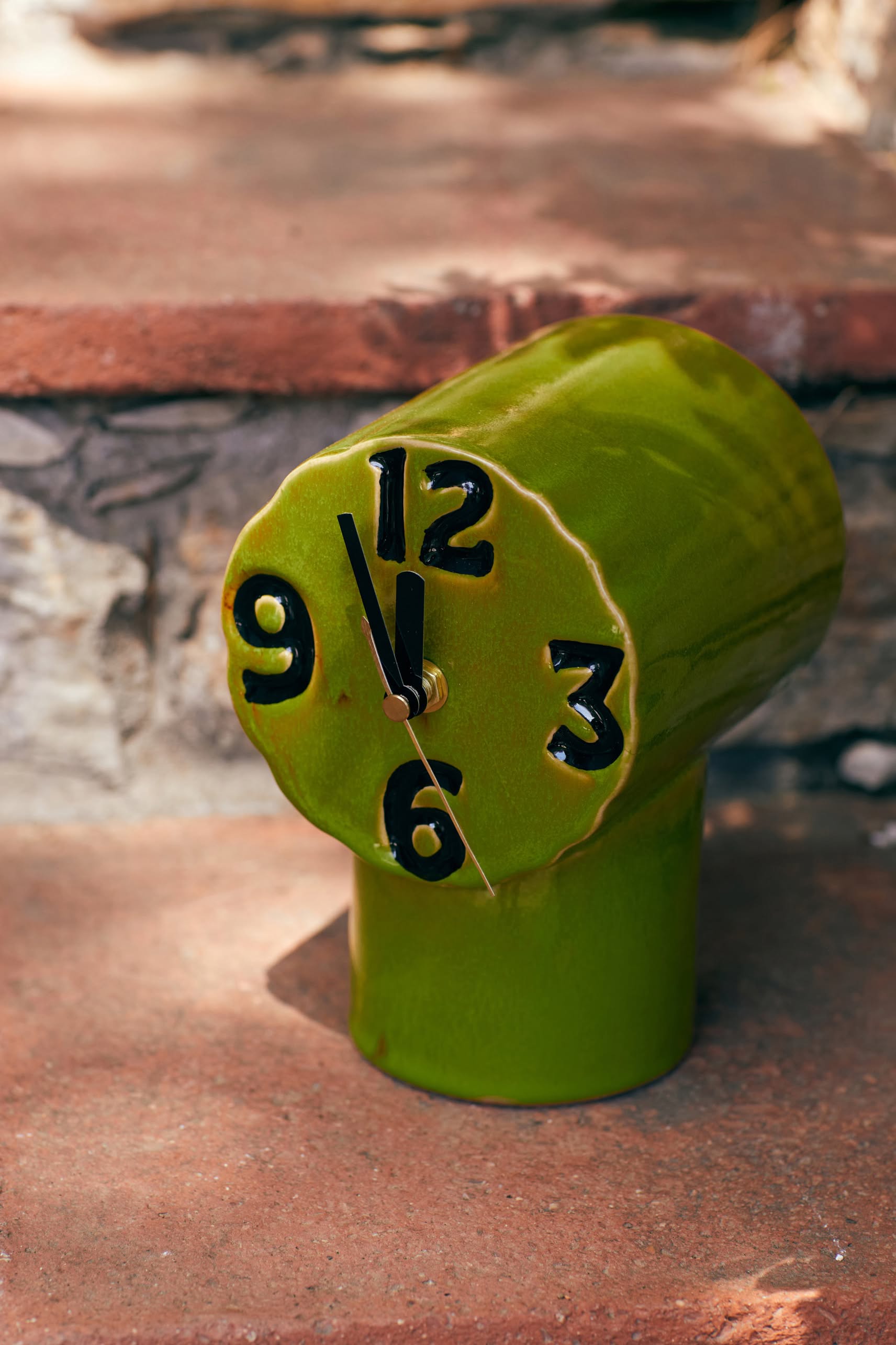 The HKLIVING Retro Ceramic Clock - Olive by HKLIVING, featuring bold black numbers and a uniquely melted vintage design, rests on an outdoor stone step, inspired by surrealist art and nostalgic style.