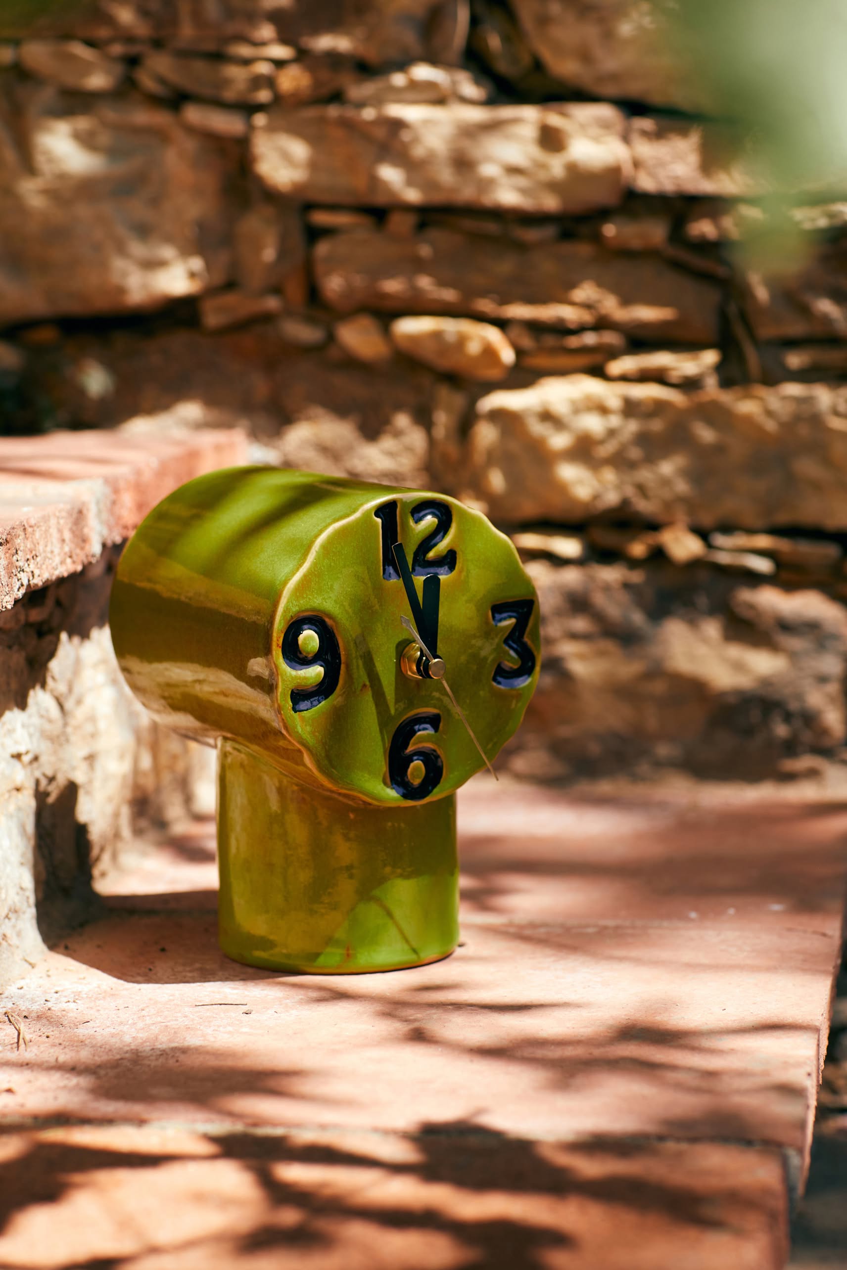 The HKLIVING Retro Ceramic Clock - Olive features bold black numbers on a melted, warped face. Sitting on a sunlit terracotta step against a stone wall, it radiates nostalgic charm and classic style from the HKLIVING brand.